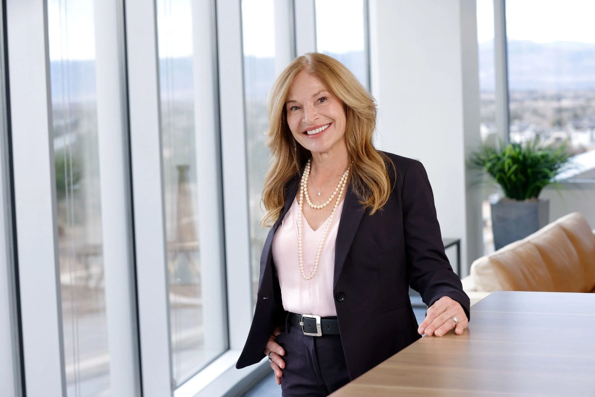 Smiling woman with blonde hair wearing a dark blazer, pink blouse, and pearl necklace, standing in a modern office with large windows and city views.