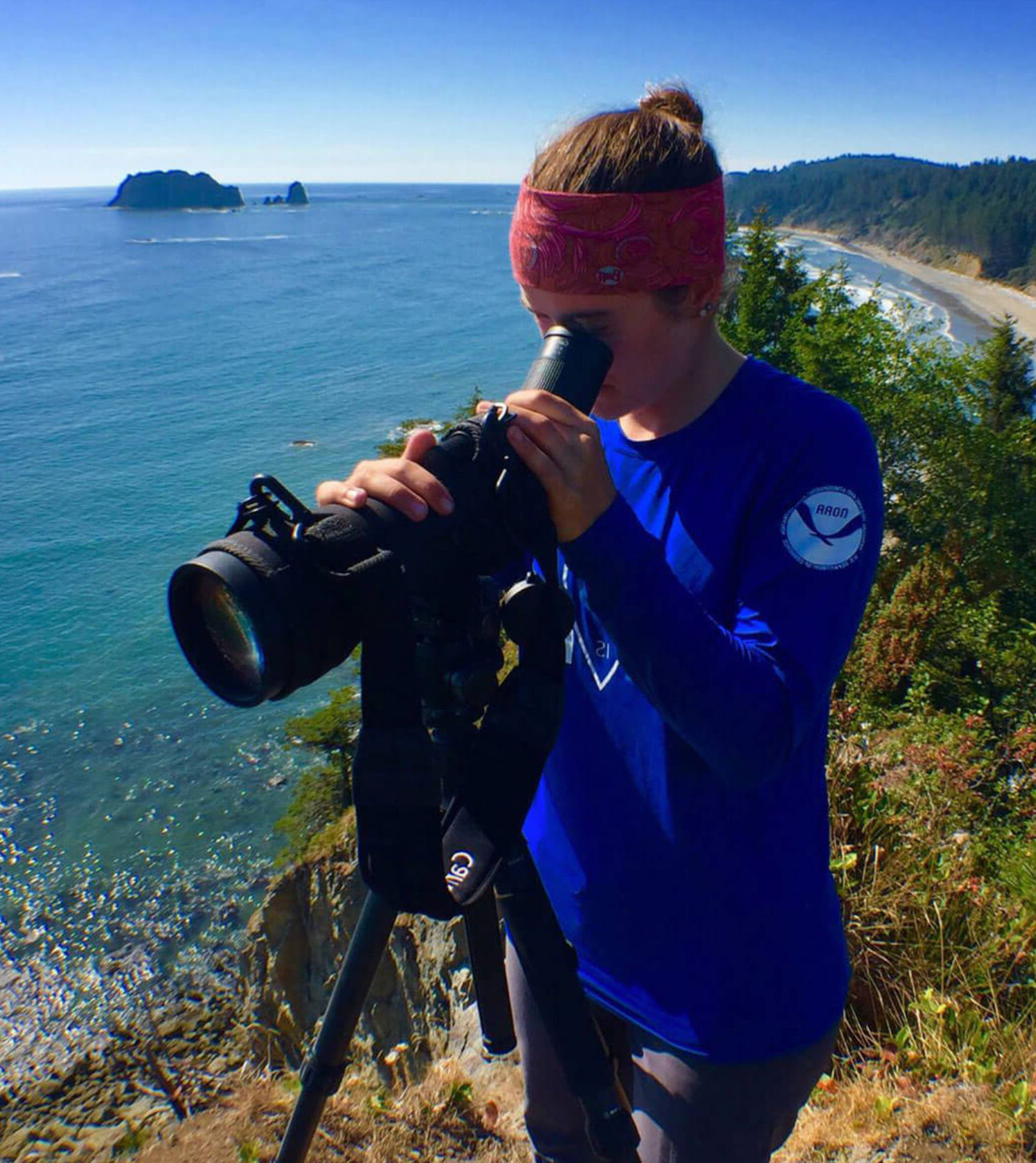 Person with orange hair, red bandana, blue long-sleeve shirt, looking through a telescope on a cliff overlooking the ocean, with islands and coastline in the background.