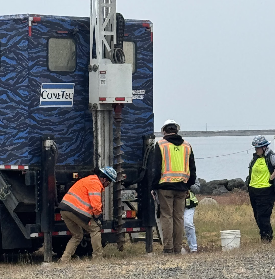 Workers and engineers wearing safety vests and helmets working near a large blue utility or maintenance truck with a crane, by a body of water and rocks.