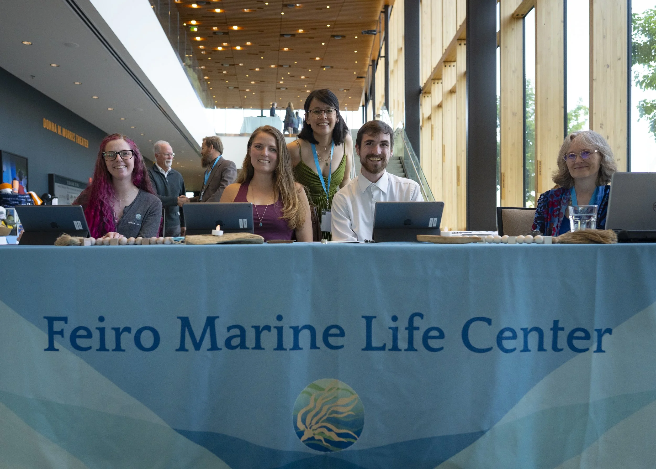 A group of five people, three women and two men, sitting and standing behind a table with a sign that reads 'Feiro Marine Life Center.' The setting appears to be an indoor exhibition or conference area with large windows and a high ceiling. The table displays electronic tablets, decorative stones, and a glass of water.