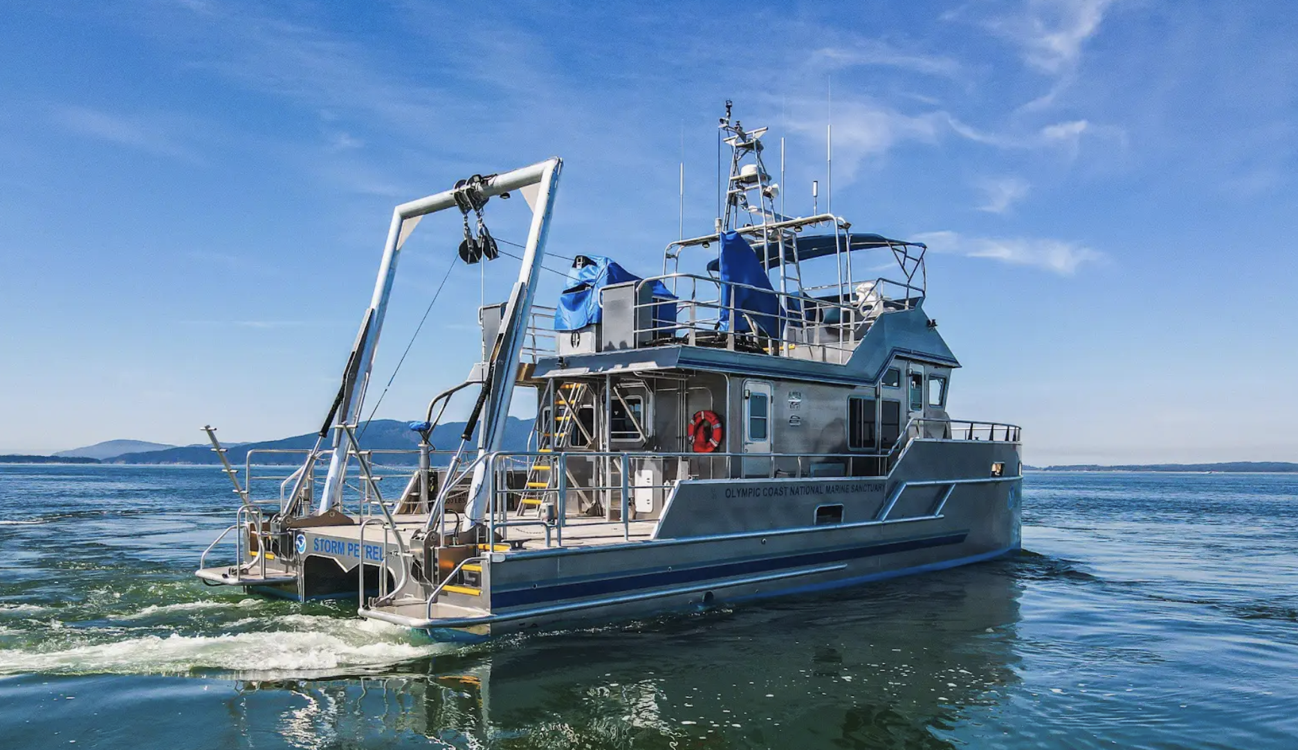A gray coast guard boat is sailing on a calm body of water under a clear blue sky, with distant land visible on the horizon.