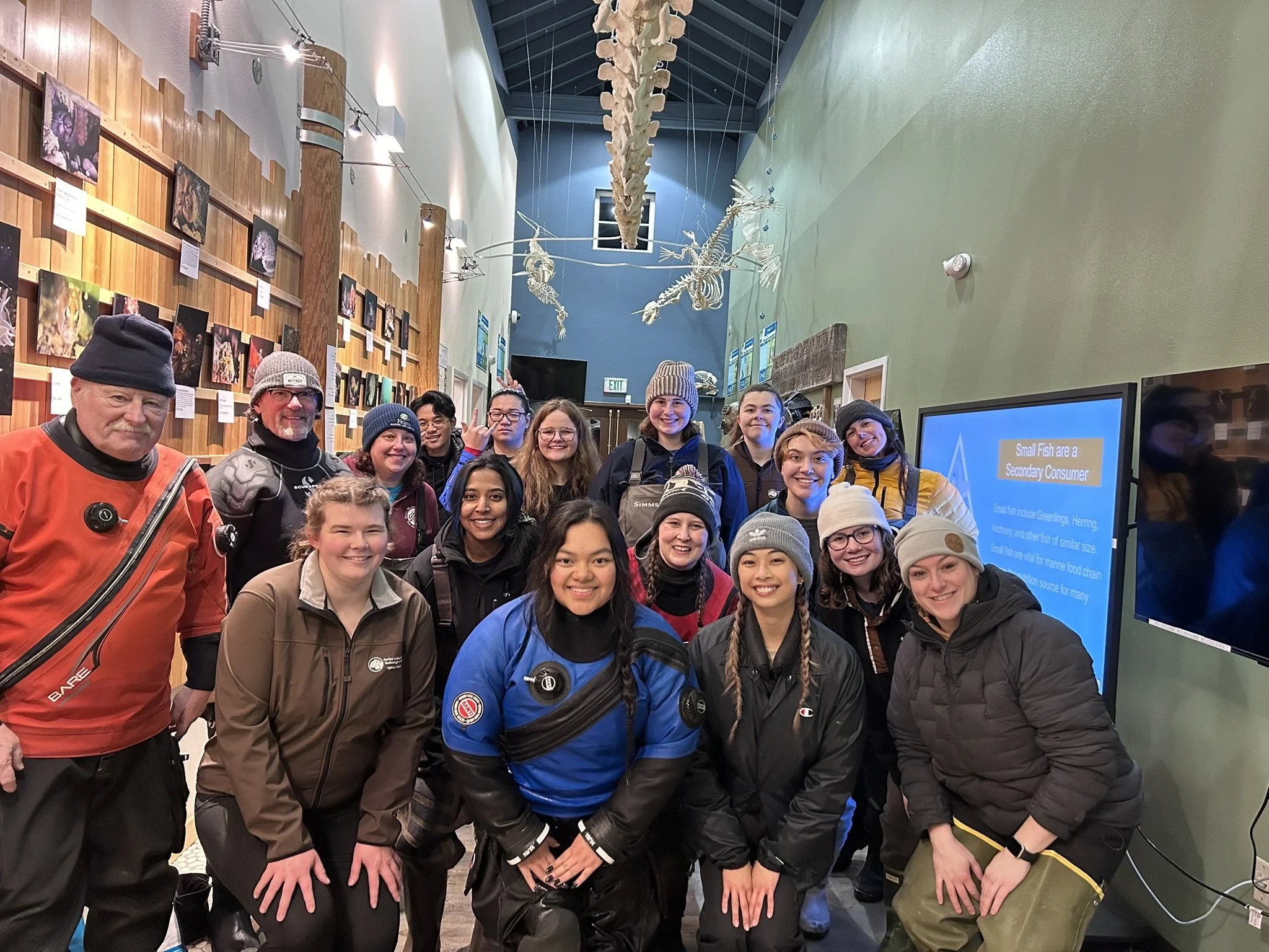 A group of diverse people gathered inside a museum, smiling at the camera, with displays of animal skeletons and photographs on the walls behind them.