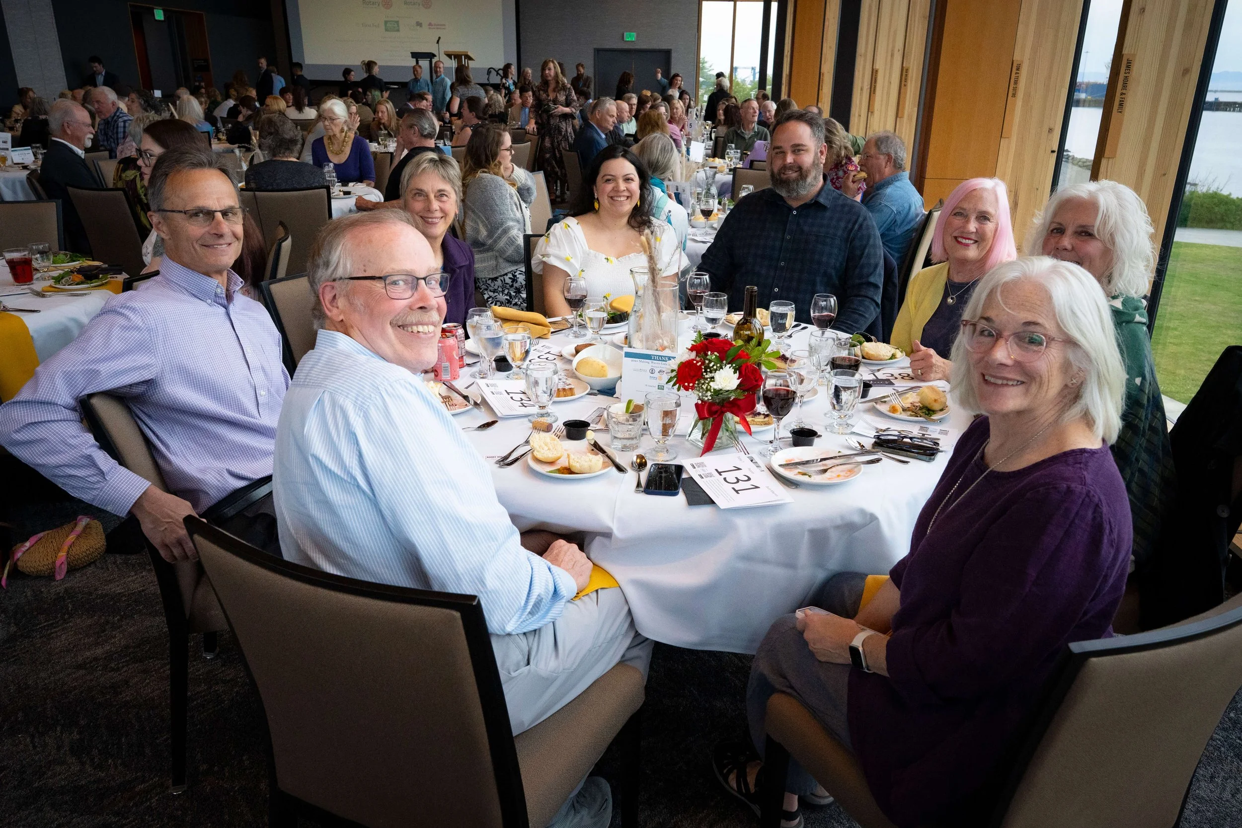 Group of people smiling and sitting around a round table at a banquet or event, with food and drink on the table, in a well-lit room with large windows showing a body of water outside.