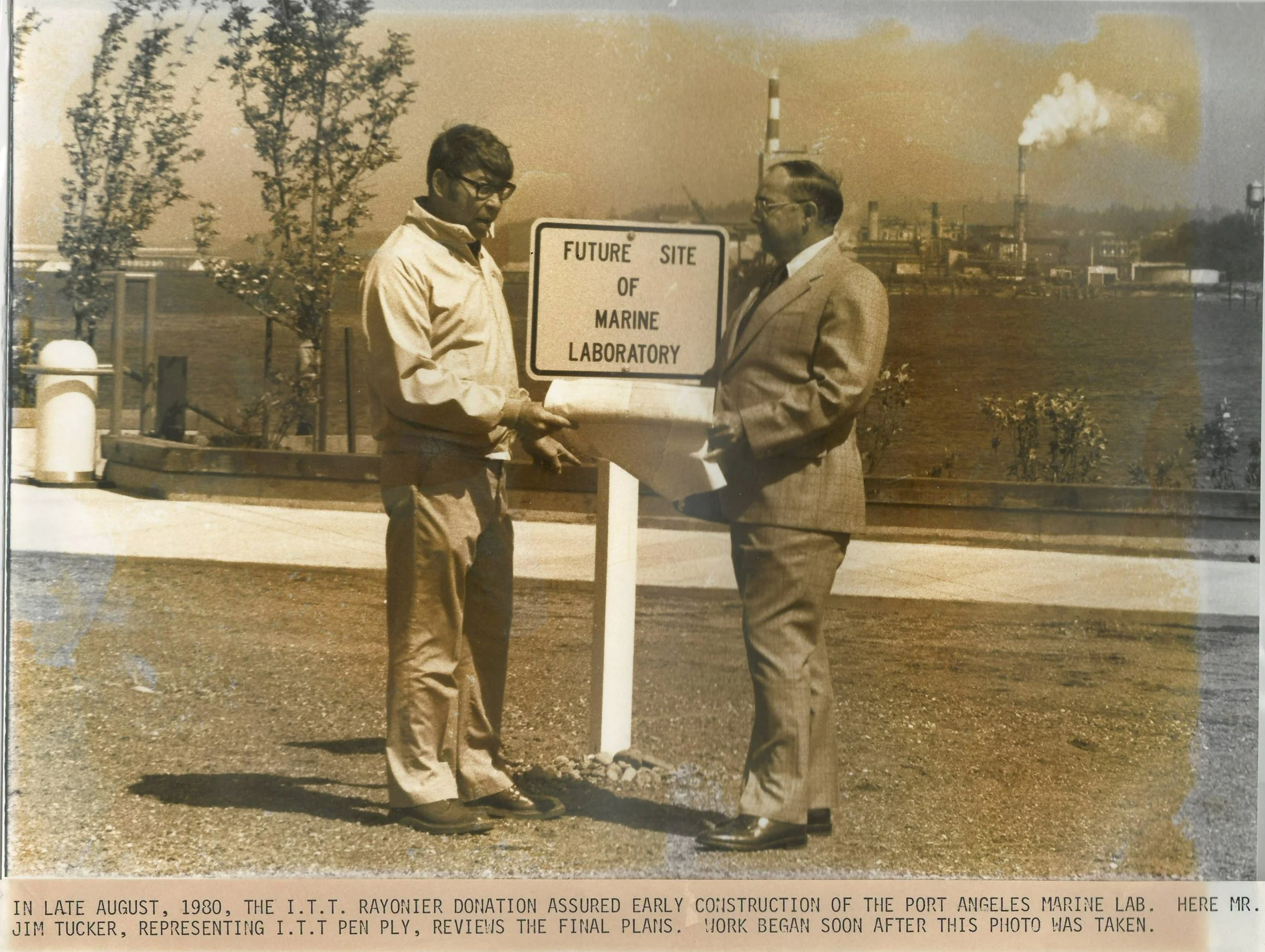 Two men standing outdoors next to a sign that reads "Future Site of Marine Laboratory," with industrial facilities in the background, during daytime in 1980.