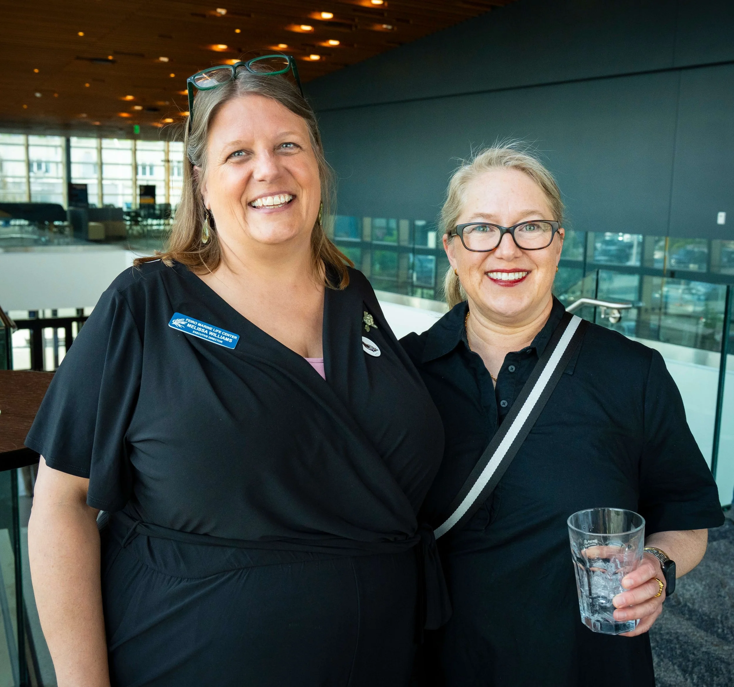 Two women smiling indoors at a modern building, one wearing glasses with a name badge, the other holding a glass of water, both dressed in black.