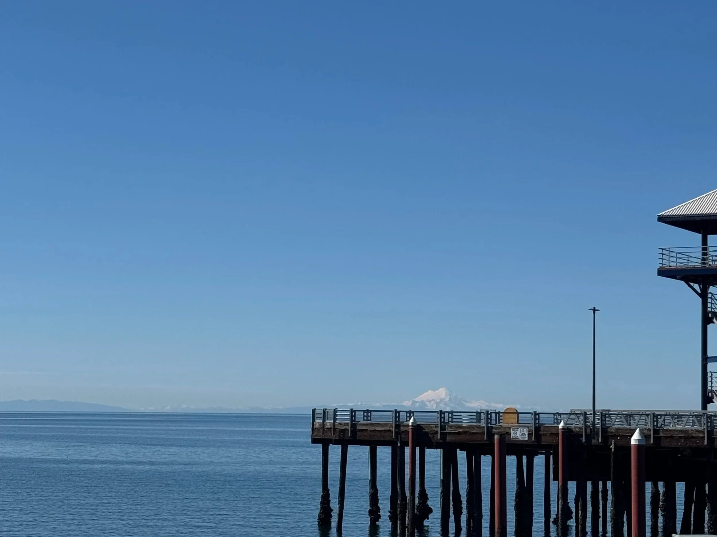Calm ocean with a pier extending into the water, a lamppost on the pier, a building with a balcony on the right, and a snow-capped mountain in the background under a clear blue sky.