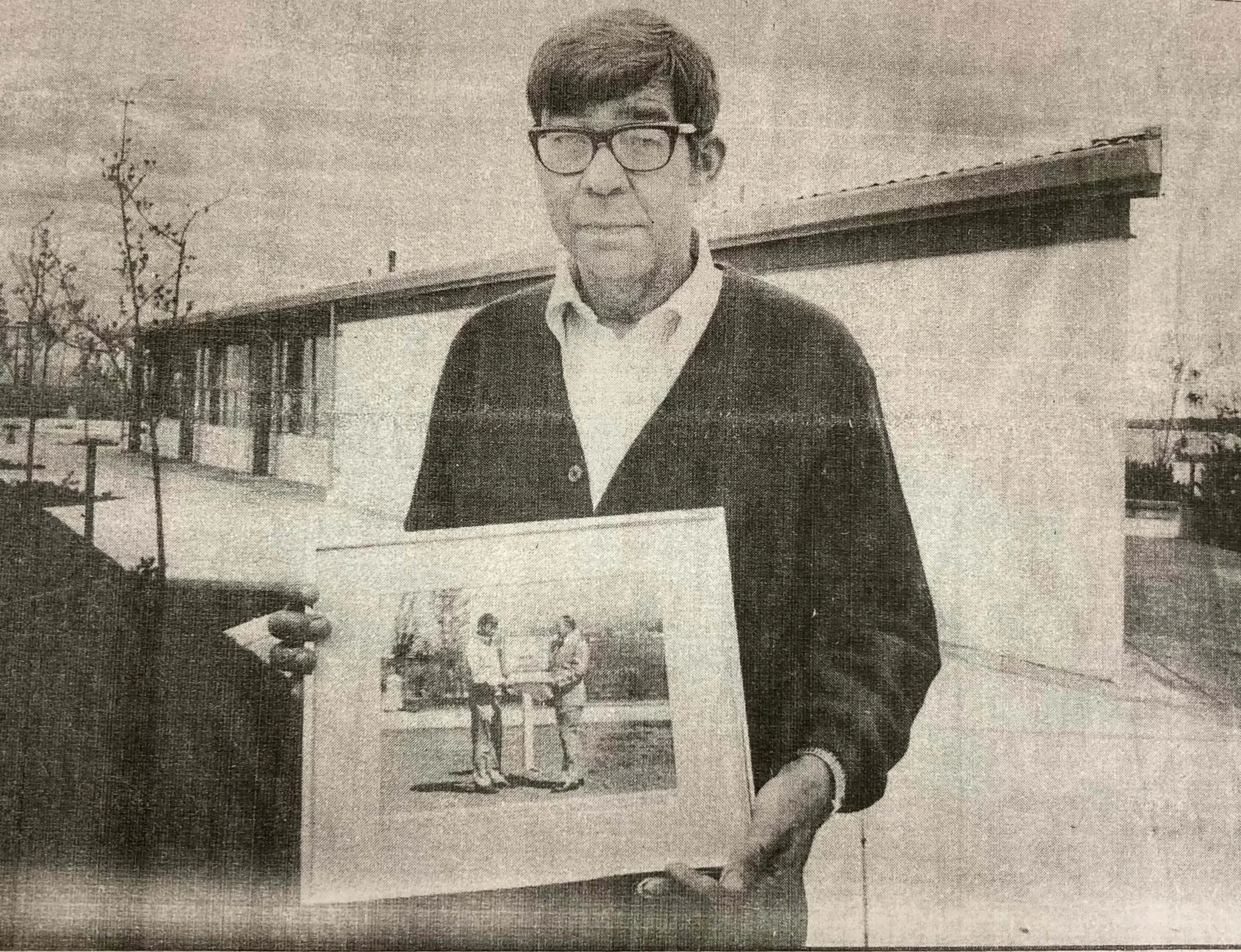 A man with glasses wearing a white shirt and dark cardigan, holding an illustration of two men in a park setting. The background shows a modern building with large windows and a clear sky.
