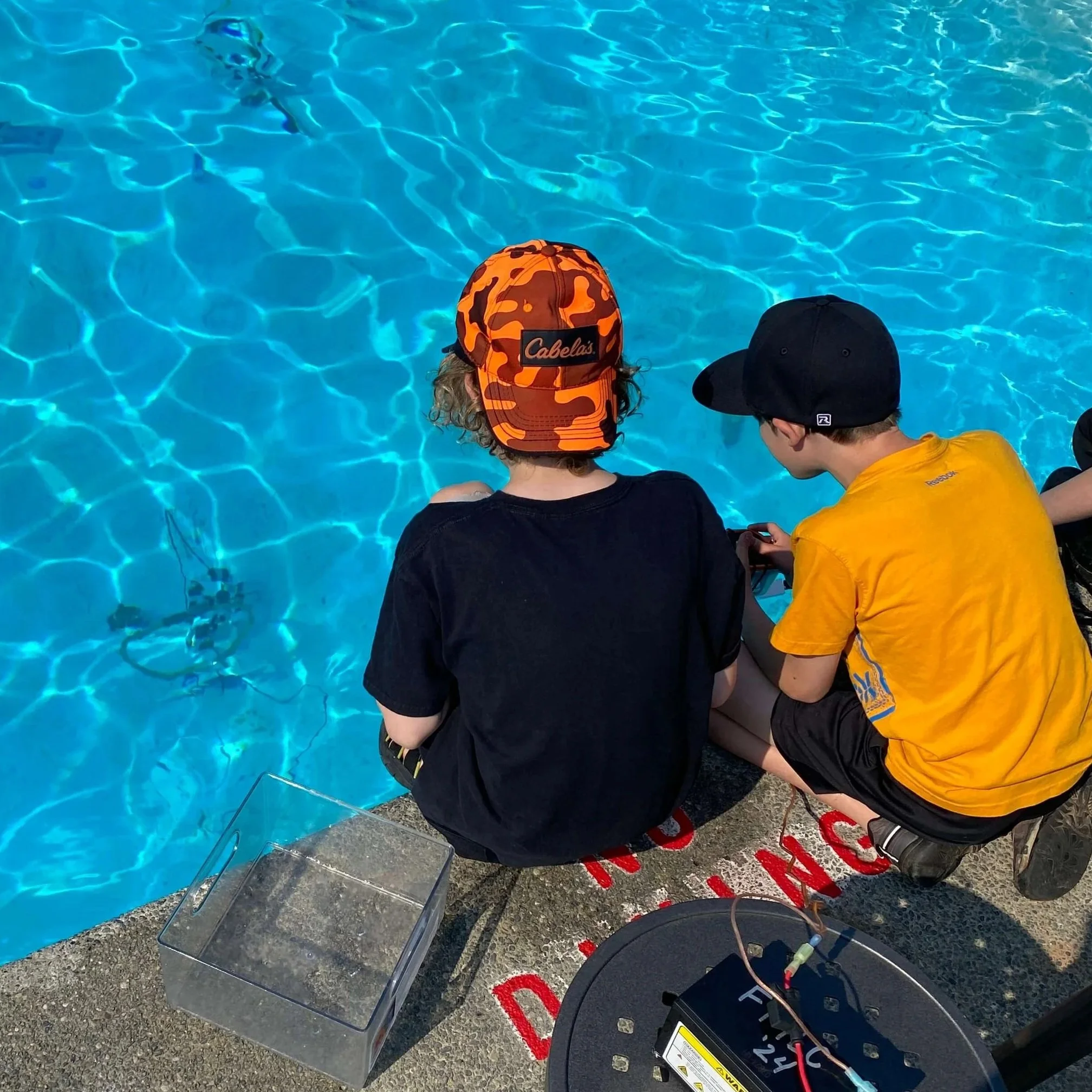 Two boys sitting by the edge of a swimming pool, one wearing an orange camouflage cap with the brand 'Cabelas' and the other wearing a black cap and yellow shirt, working with electronic equipment.