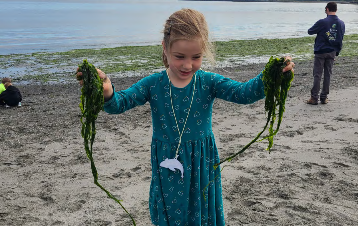 A young girl on a beach holding up two large pieces of seaweed, with a body of water and trees in the background.