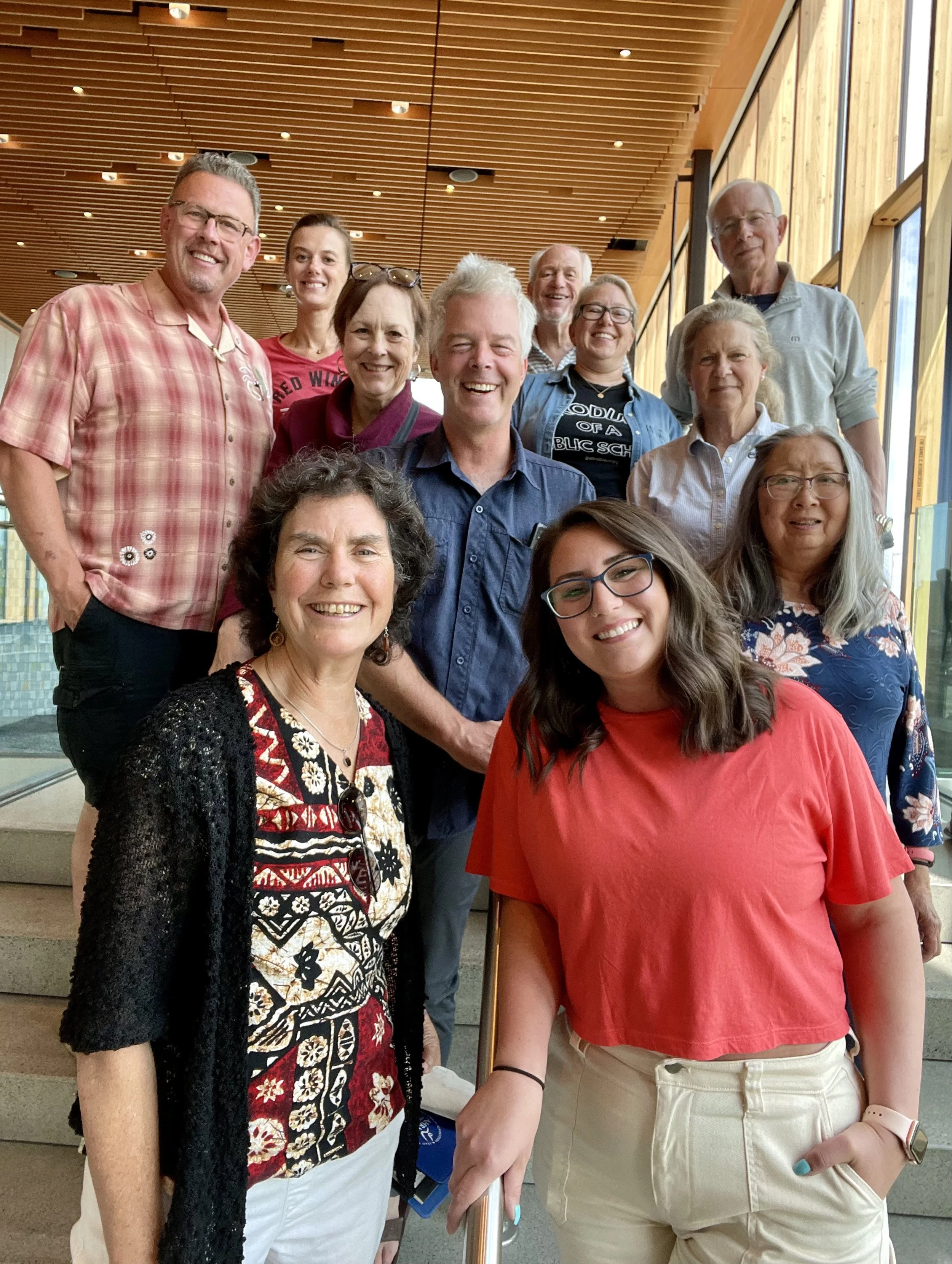 Group of people smiling for a photo indoors, standing on stairs near large windows with wooden paneling ceiling.