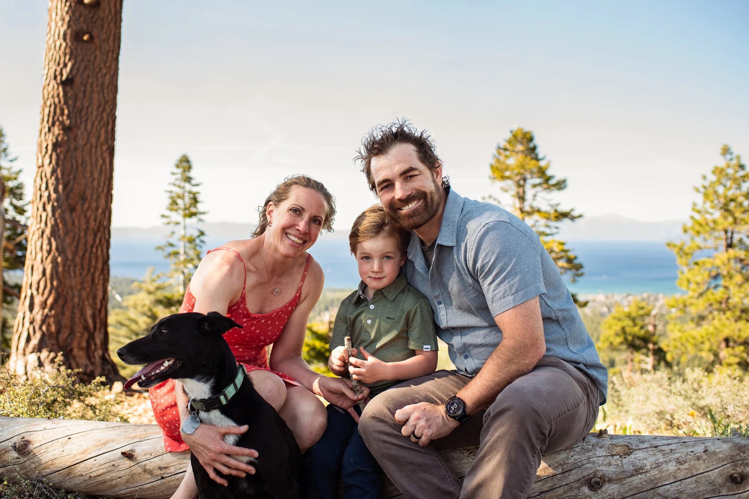 Family of three with their dog outdoors in a forested area with mountains and lake in the background, smiling for the photo.