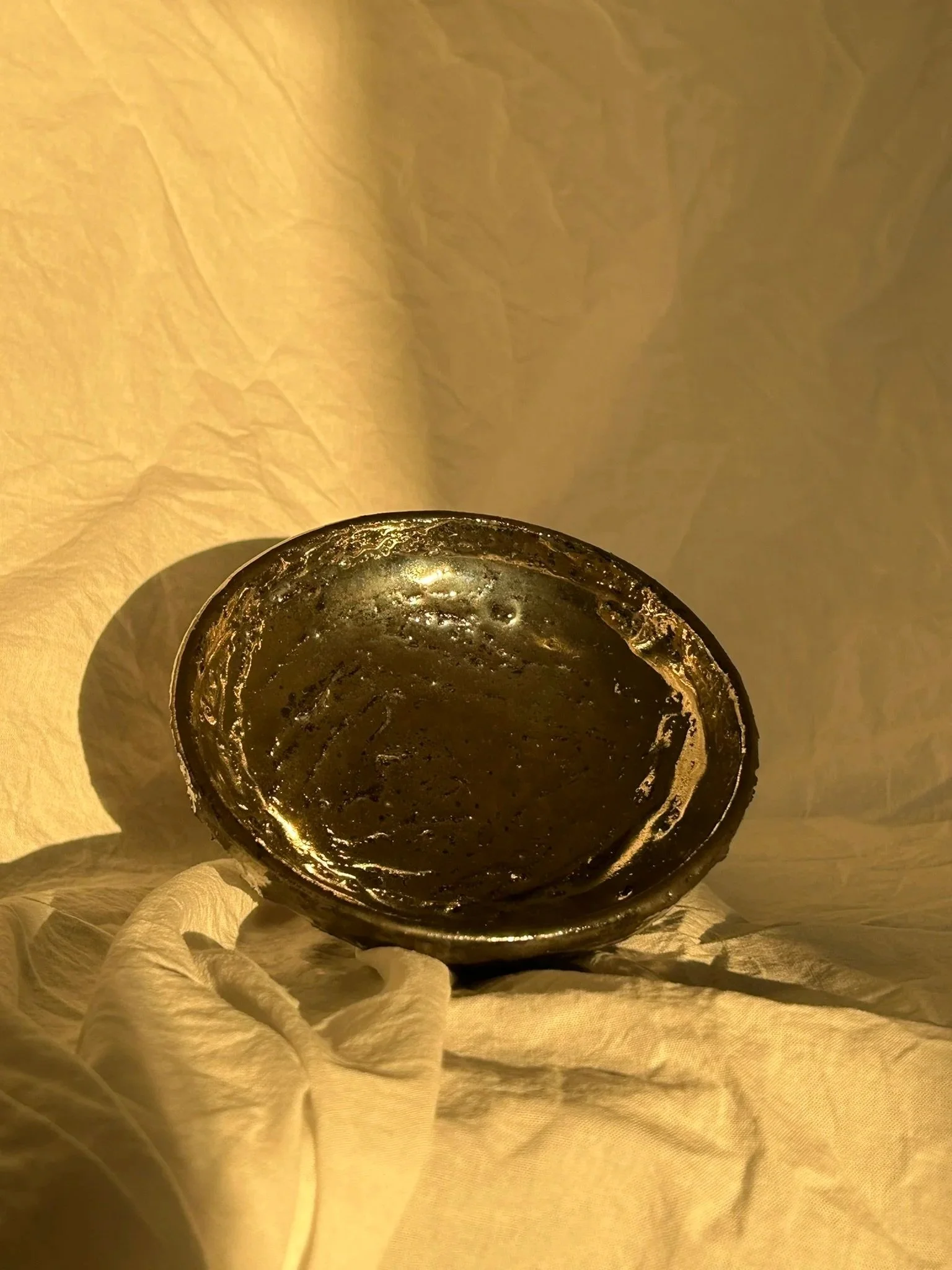 A dark, shiny, round-bottomed bowl or dish held by a person's hand wearing a white cloth glove, set against a yellowish background.