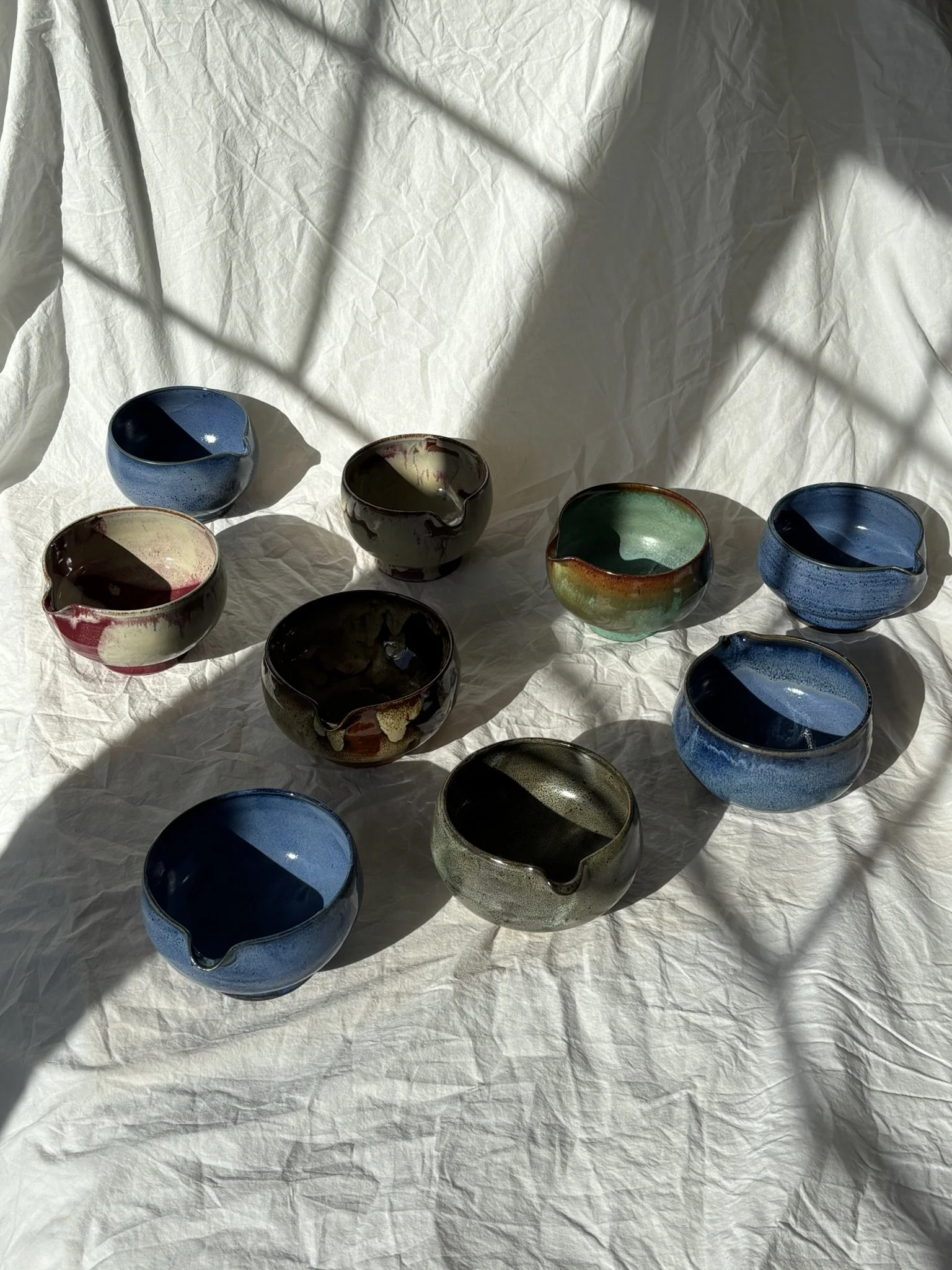A collection of ten ceramic bowls with pouring spouts, arranged on a crinkled white fabric. The bowls have various glazed colors including blue, black, green, and multicolored, and are casting shadows from sunlight and window shadows.