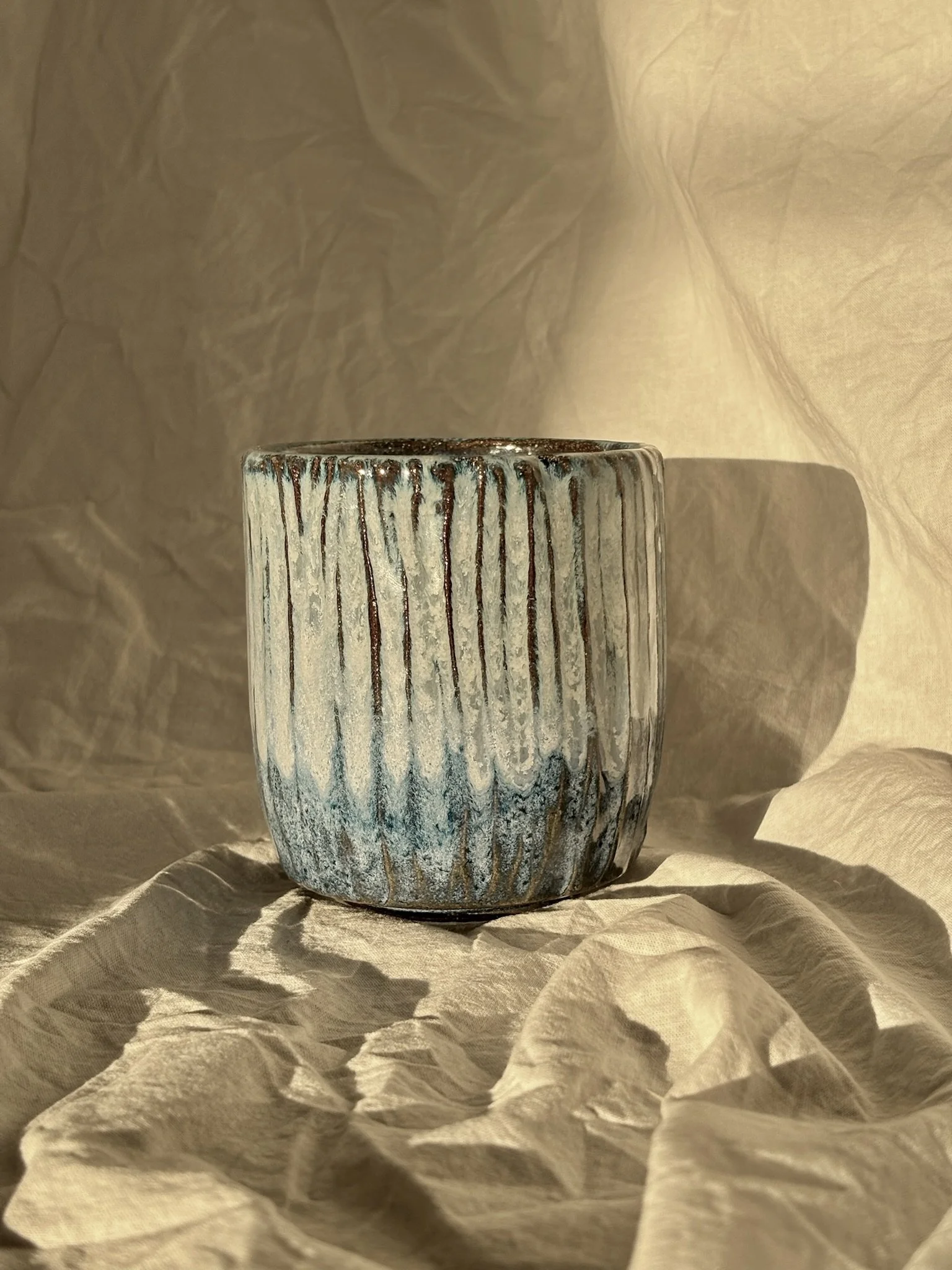 A ceramic cup with vertical ridges and a textured glaze, sitting on crumpled beige fabric with a shadow cast to the right, illuminated by warm, soft lighting.