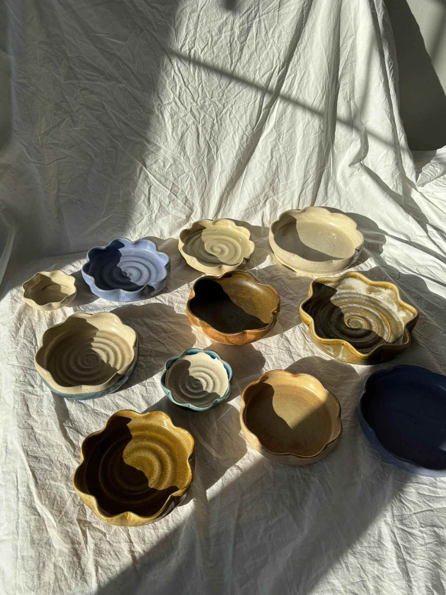 Various small, colorful, flower-shaped ceramic bowls arranged on a white cloth surface with sunlight casting shadows.