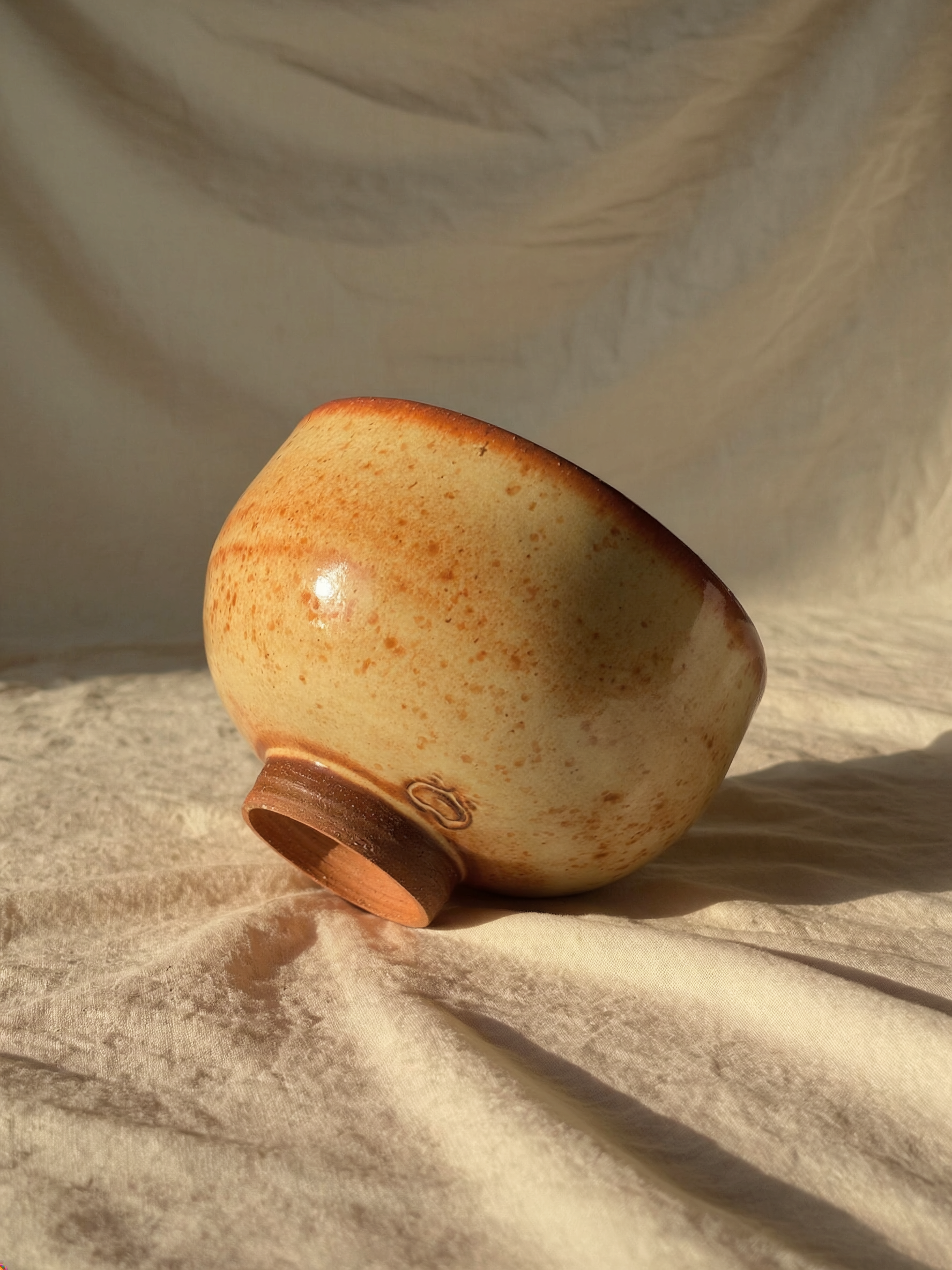 A ceramic vase with a speckled beige and brown pattern, lying on its side on a textured beige fabric surface, with soft lighting and shadows