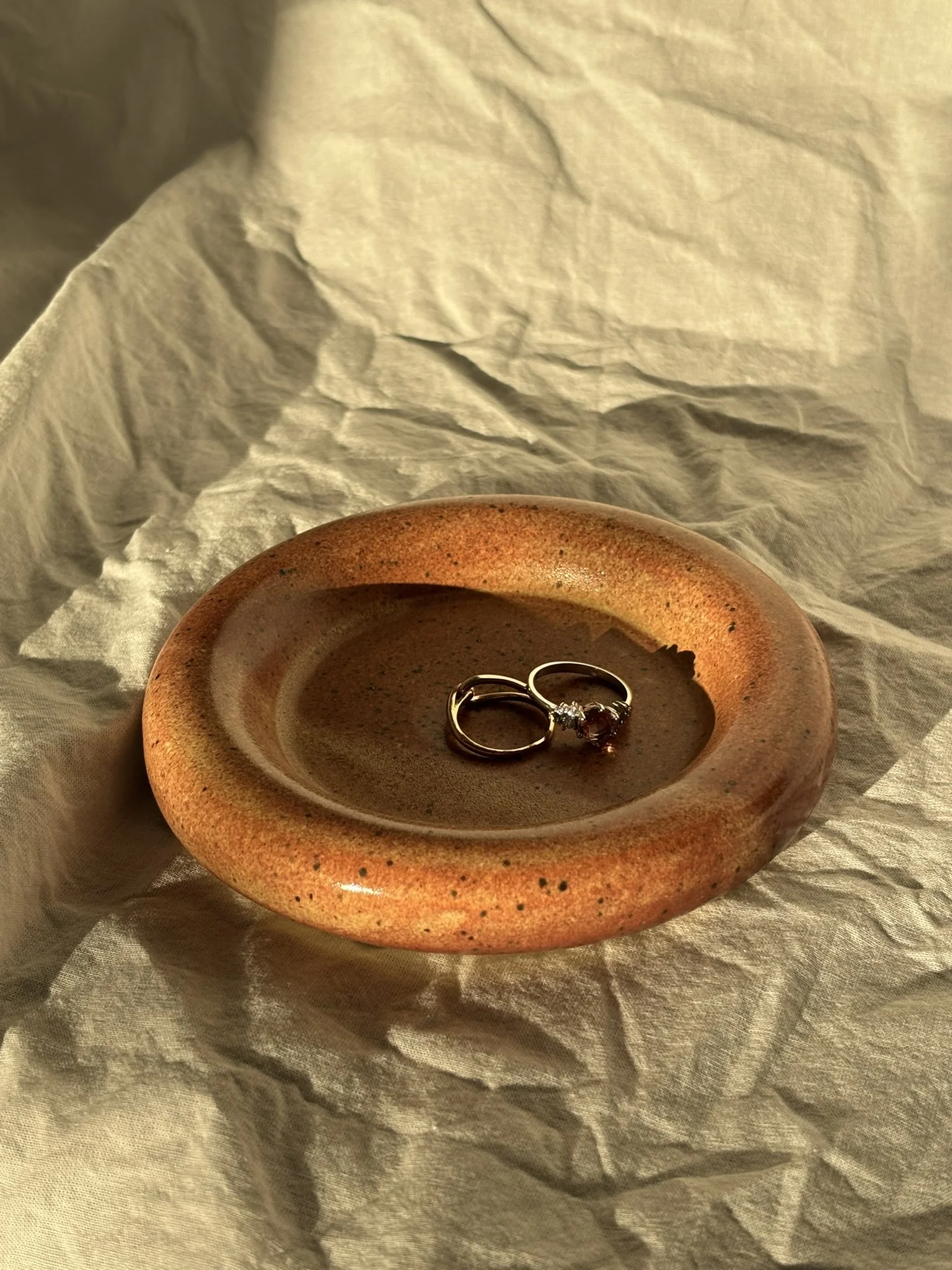 A small, round, textured ceramic dish with a shallow indentation, gentle speckles and a warm earthy color, contains a silver ring with a gemstone inside.