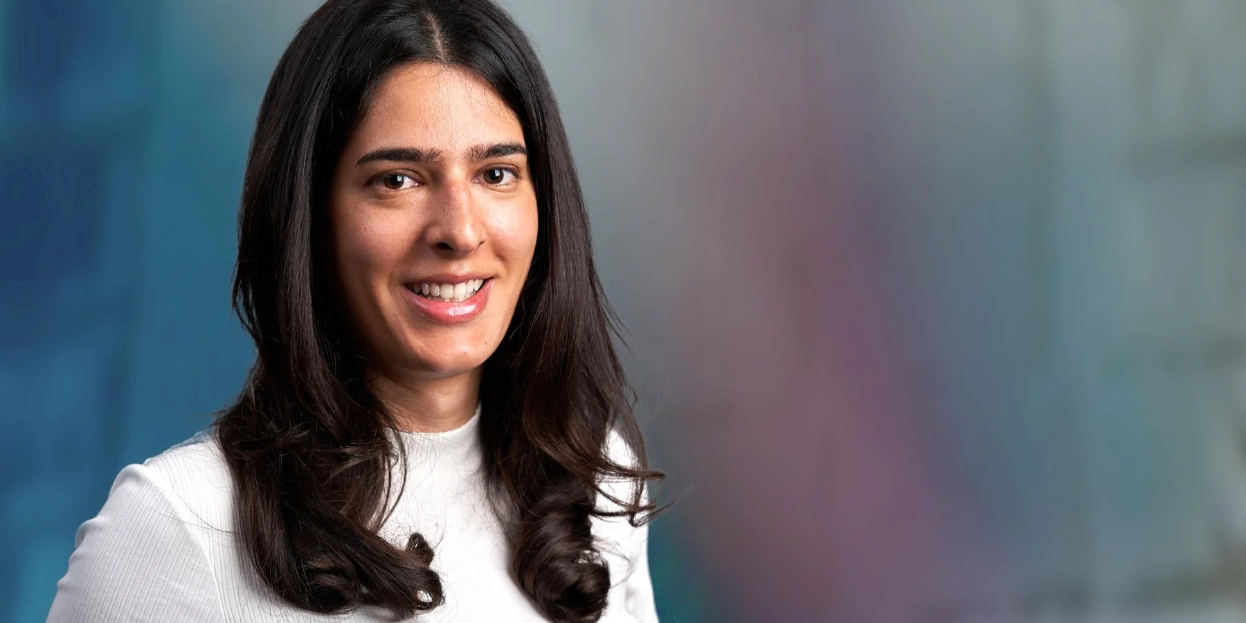 A woman with long dark hair smiling, wearing a white top, with a blurred colorful background.