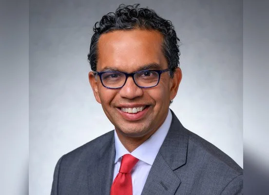 Professional portrait of a smiling man with curly black hair, wearing glasses, a gray suit, white shirt, and red tie against a gray background.