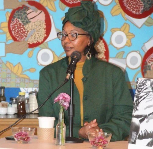 Woman wearing green jacket and headwrap speaking into a microphone at a table with flower vases and bowls, colorful patterned wallpaper in the background.