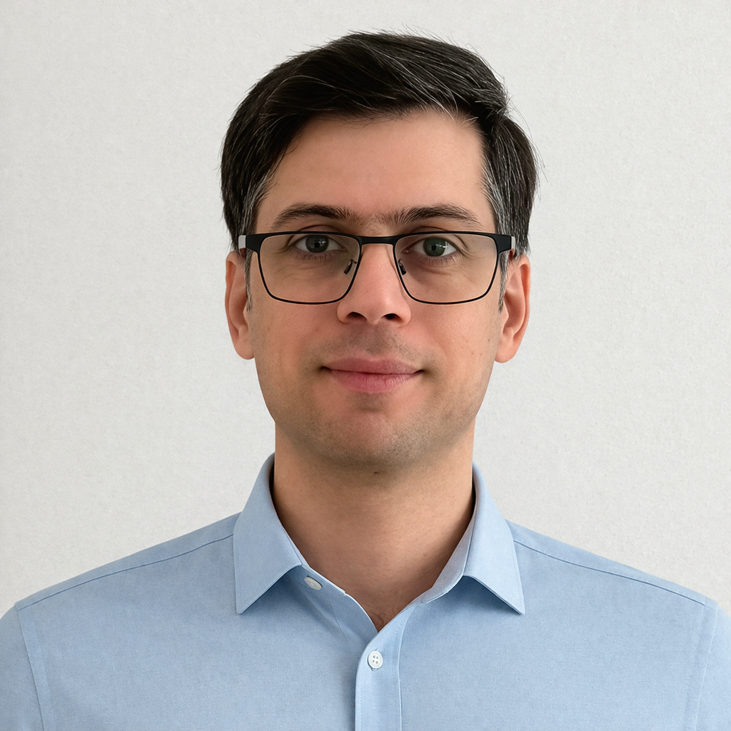 A man with short dark hair, wearing glasses and a light blue dress shirt, standing against a plain white wall.