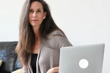 A woman with long brown hair sitting on a black couch with a laptop in front of her, holding a smartphone.