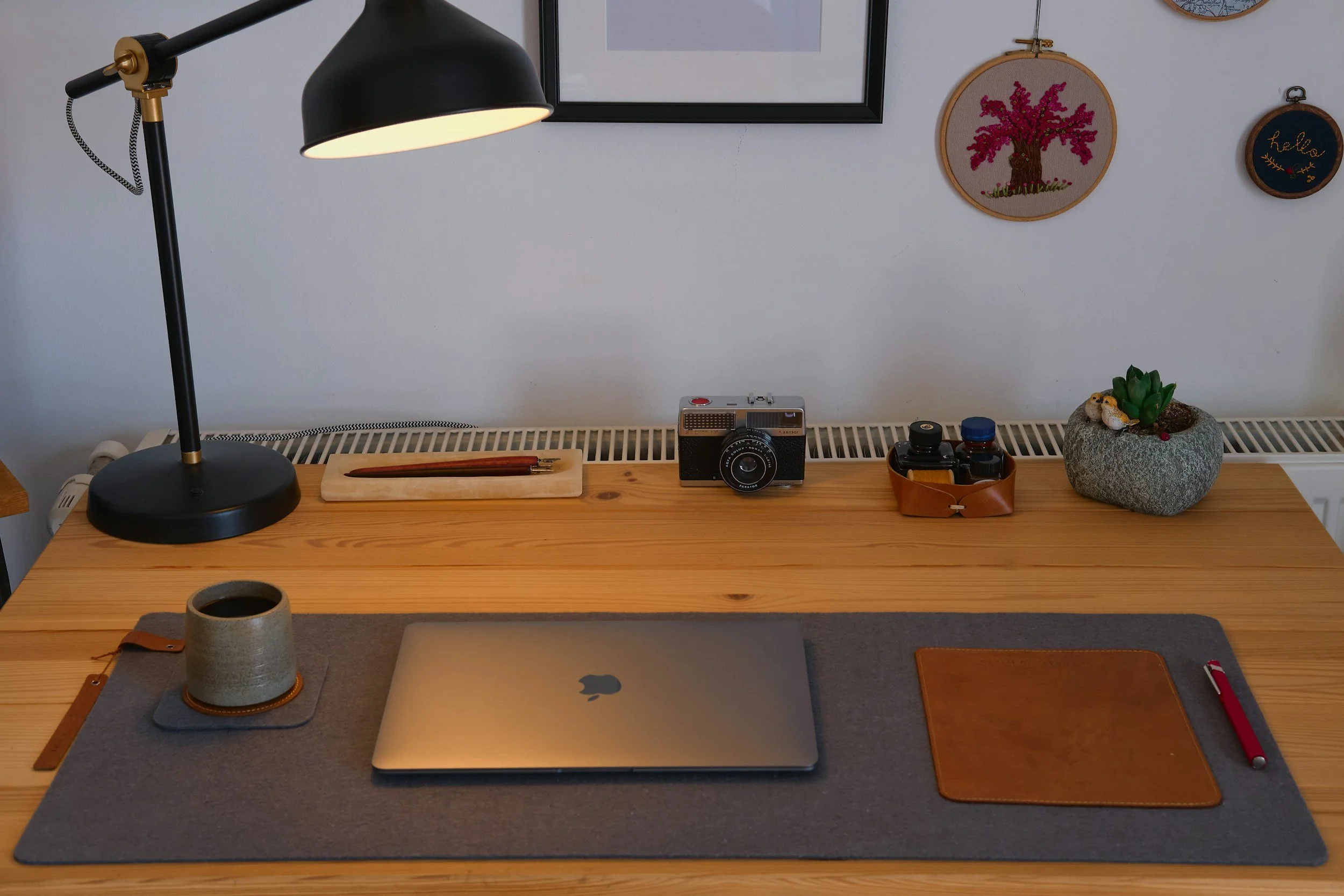A tidy workspace with a wooden desk, a black desk lamp turned on, a closed MacBook, a ceramic cup on a coaster, a brown leather patch, a red pen, a vintage camera, a small wooden container with bottles, and a decorative plant in a rock-like pot. Hanging on the wall are framed pictures and embroidered wall art.