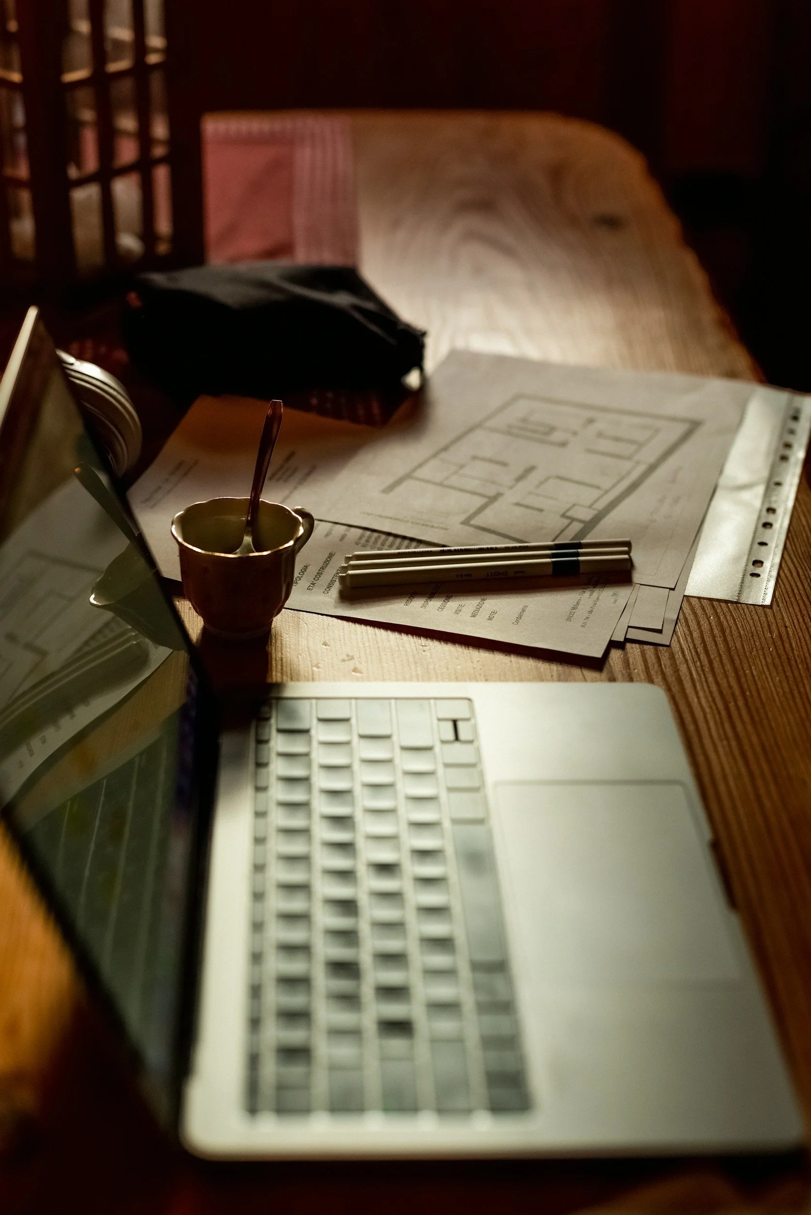 A wooden table with architectural plans, pens, a small cup with a spoon, a closed laptop, and a black pouch, illuminated by warm lighting.