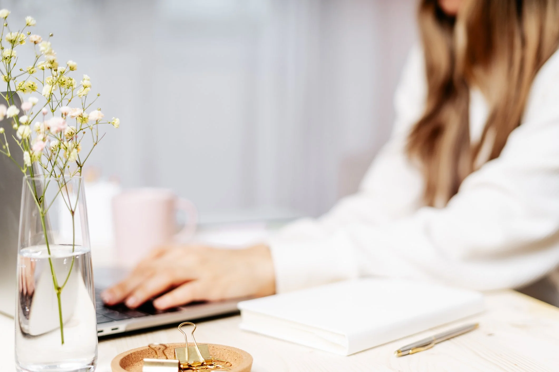 A person working on a laptop at a desk with a glass vase of flowers, a notebook, a pen, and a small dish with gold paper clips.