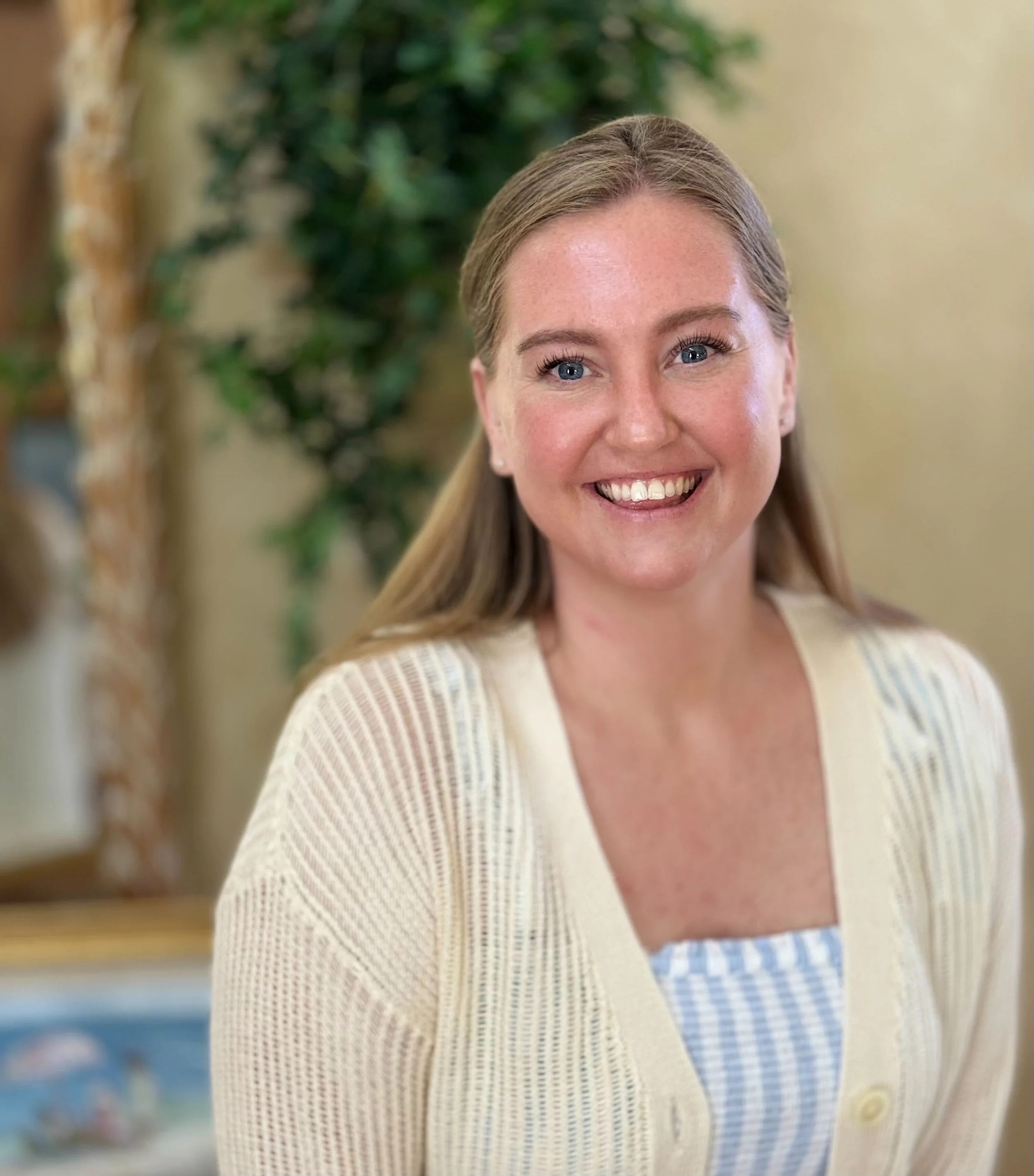 A smiling woman with long blonde hair, wearing a cream-colored cardigan and a blue and white striped top, standing indoors with a blurred green plant and yellow wall in the background.