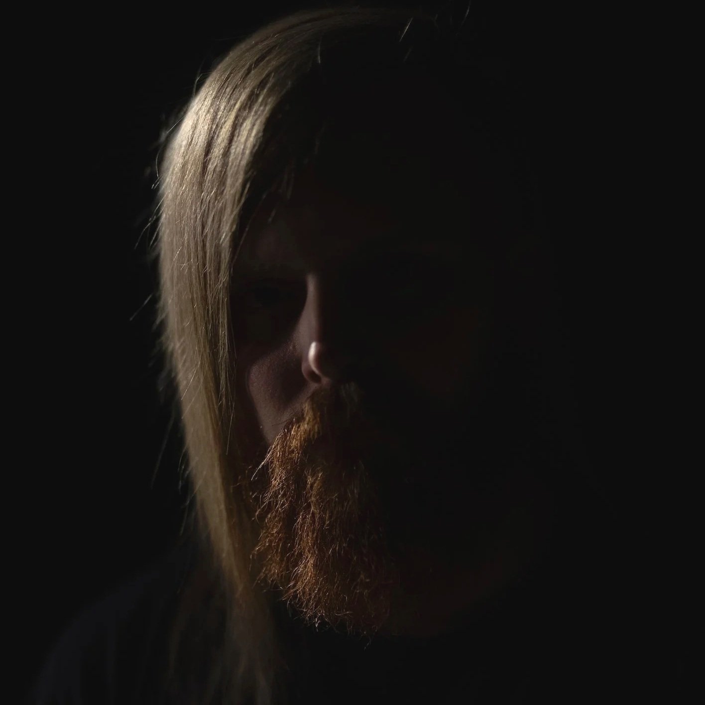 Portrait of a person with long hair and a beard, partially illuminated against a dark background.