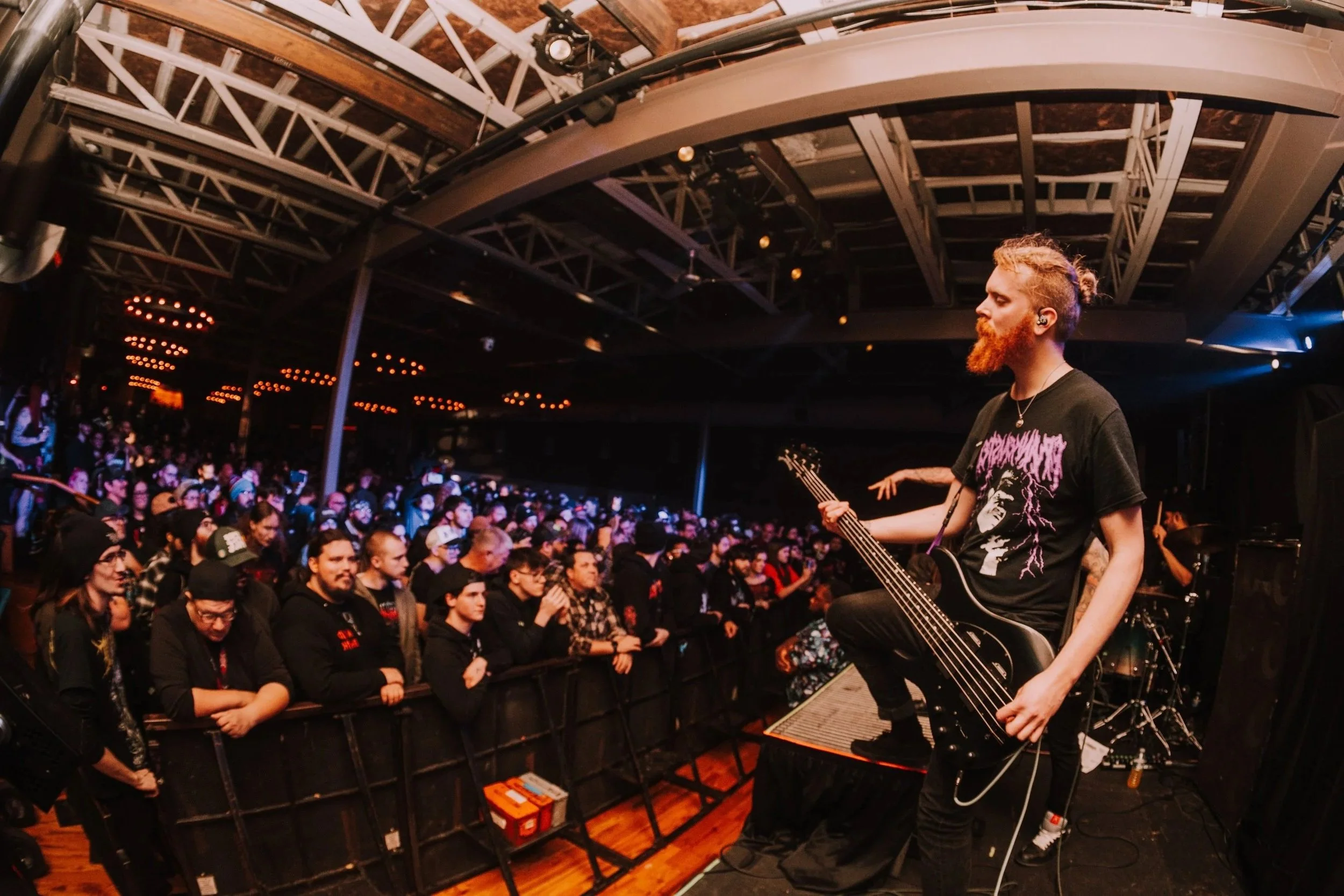 A musician with a red beard and a black graphic T-shirt playing an electric bass guitar on stage during a concert, facing the audience in a crowded venue with dim lighting and decorative ceiling lights.
