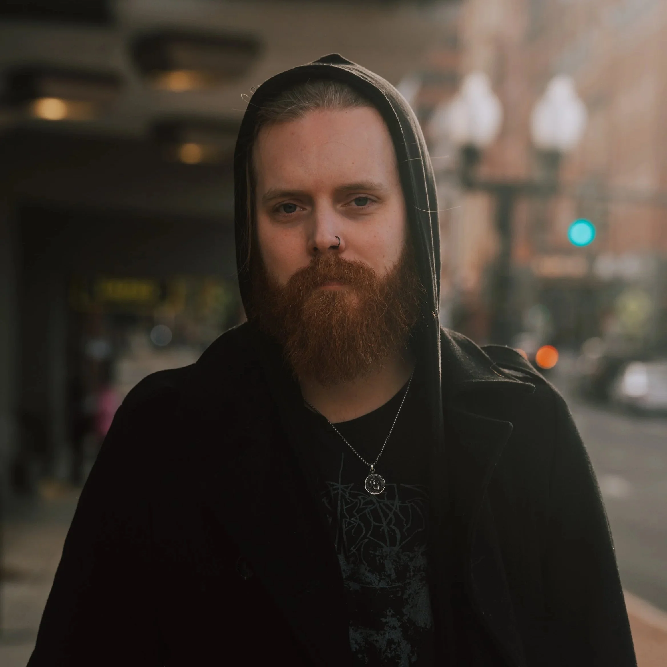 A man with a beard, wearing a black hoodie with the hood up, standing outdoors on a city street during sunset, with blurred city lights and buildings in the background.