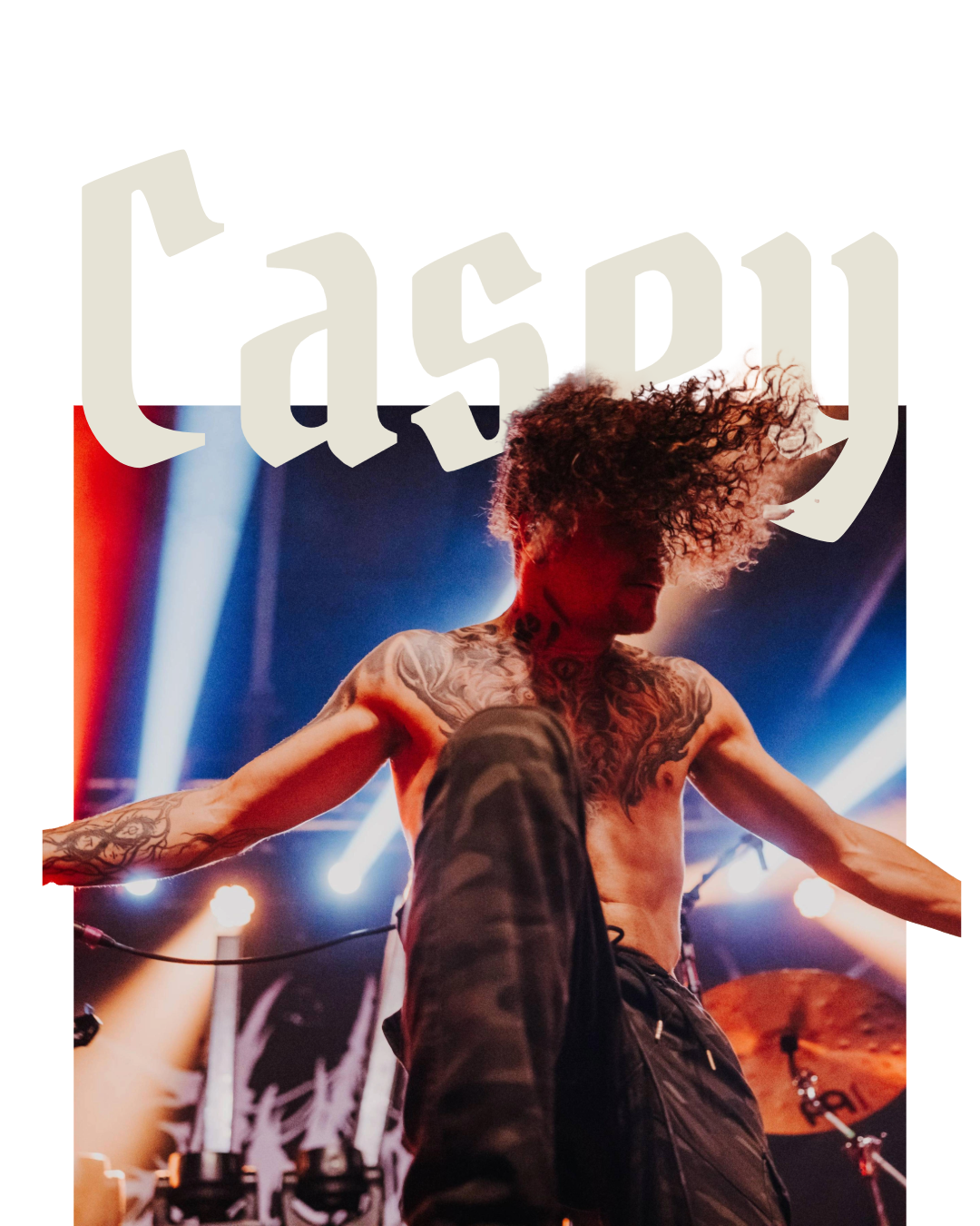 A shirtless male performer with tattoos and curly hair dancing on stage with colorful lights and drums in the background.