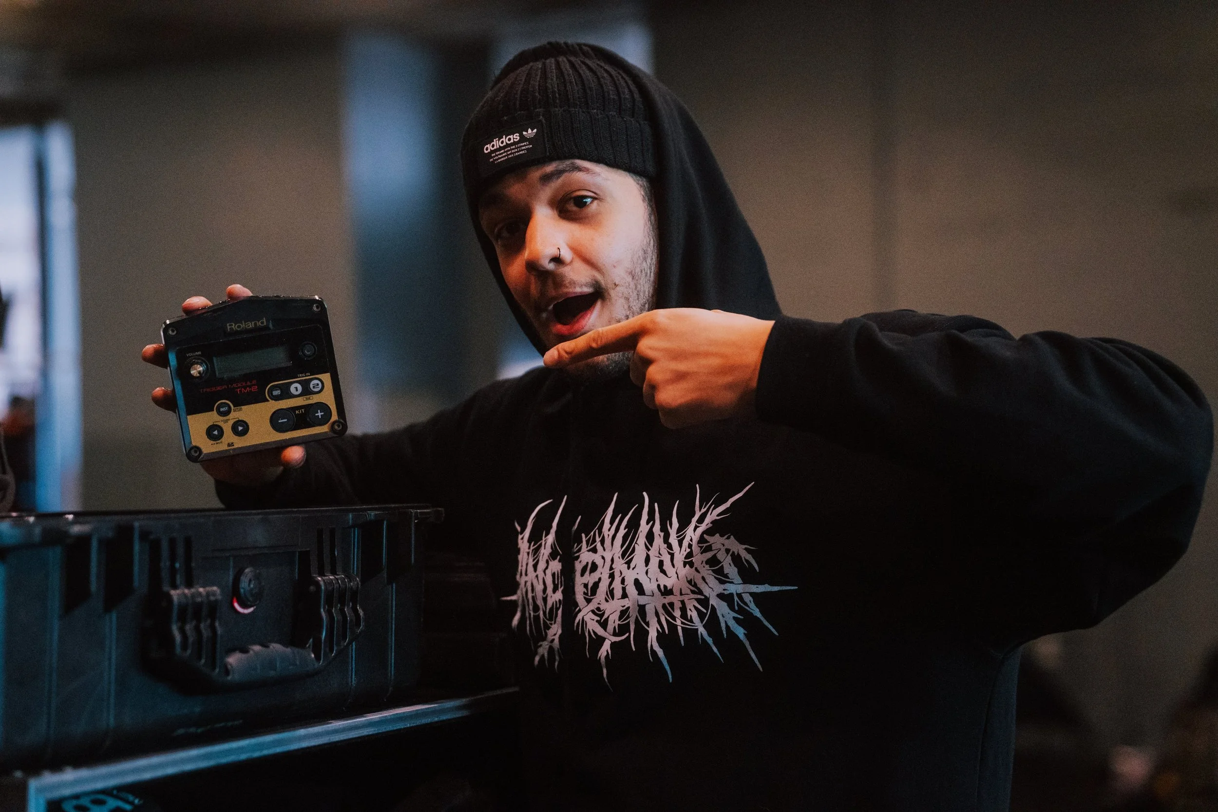 Young man in black hoodie and beanie pointing at a musical equipment device in a dimly lit room, smiling.