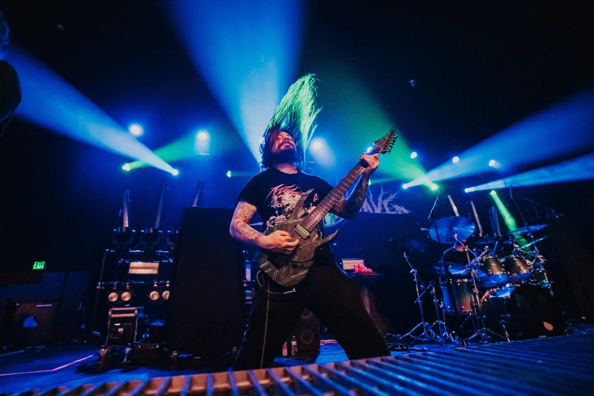 Guitarist with long hair and tattoos playing electric guitar on stage with colorful lights and a drum kit in the background.