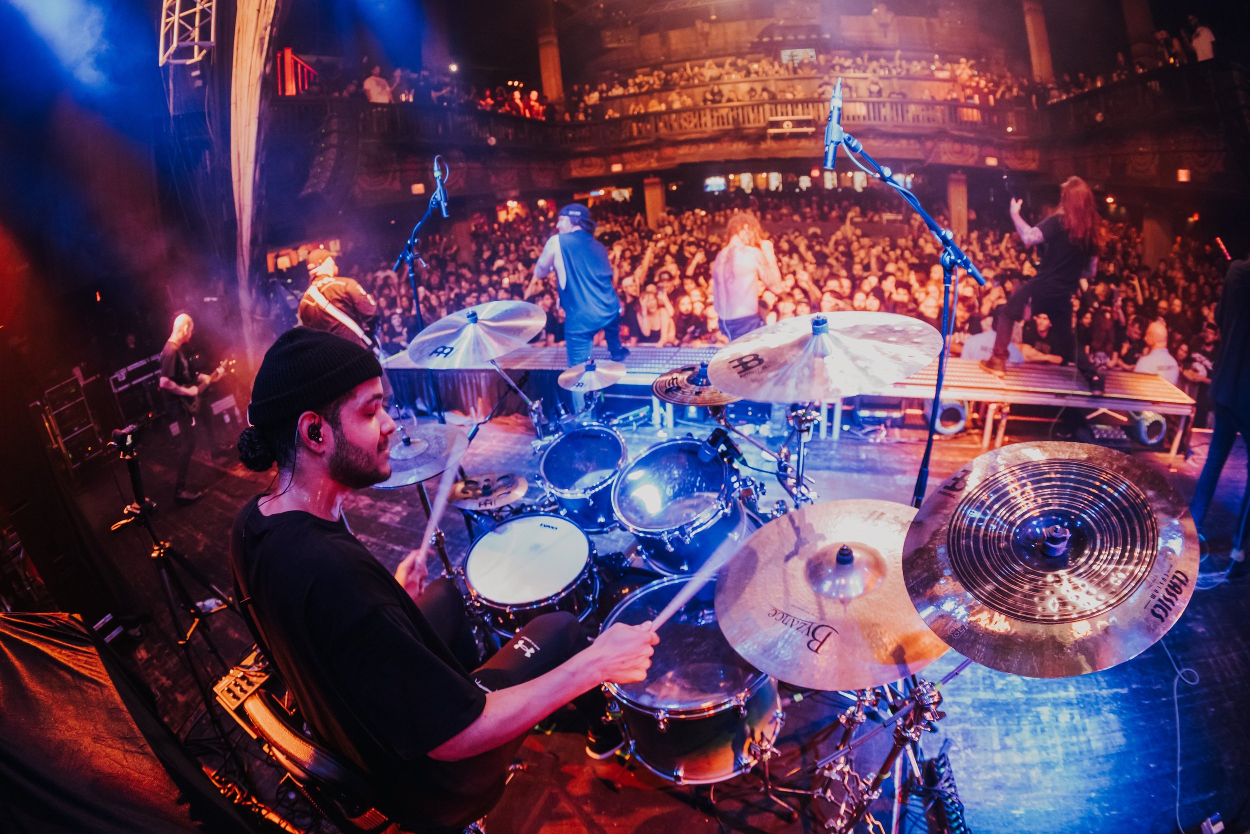 Stage view from behind a drummer performing with a band, with a large audience in the background, on stage with microphones, instruments, and performers.