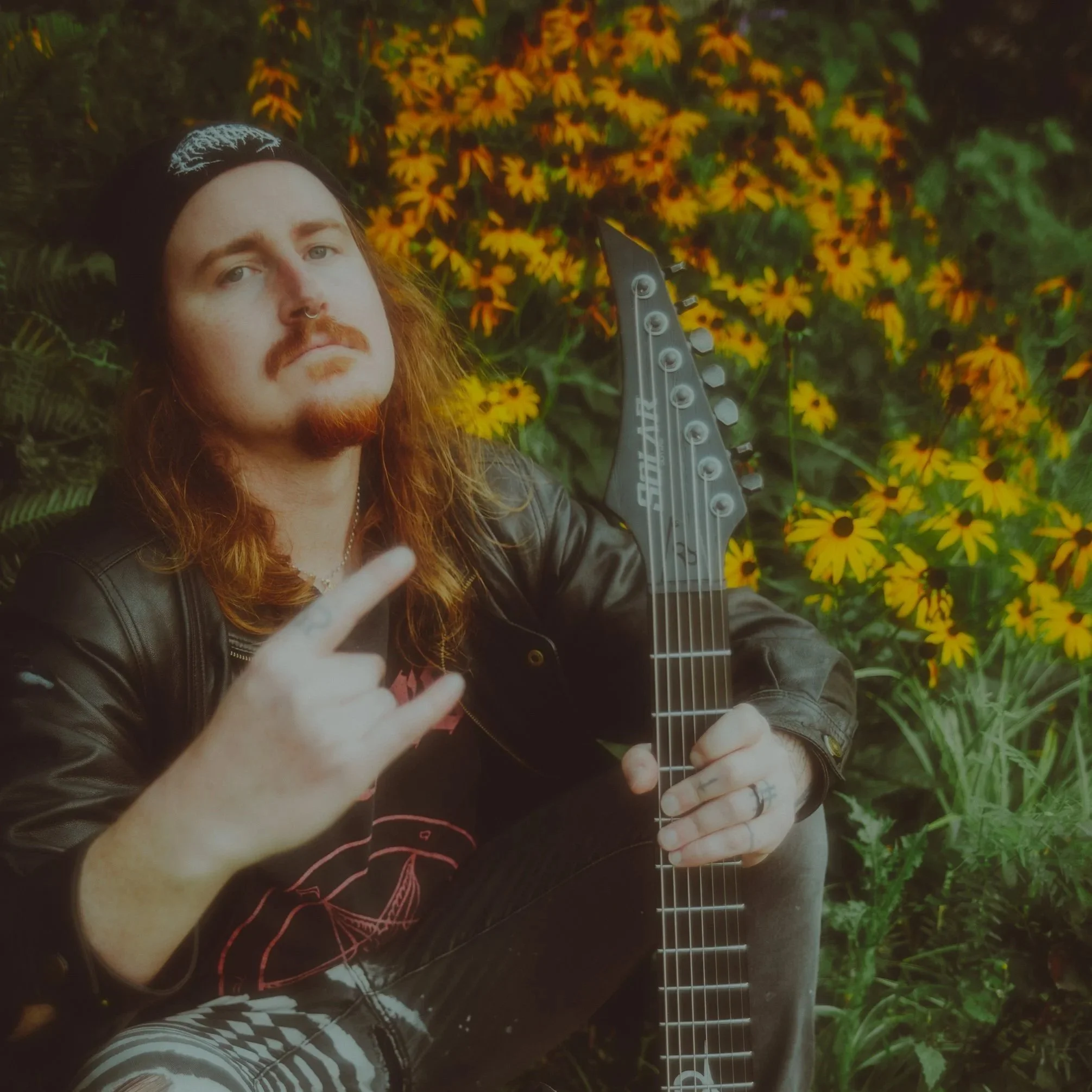 A man with long red hair, a goatee, and a nose ring, wearing a black beanie and leather jacket, holding an electric guitar, displaying a rock hand gesture, sitting in front of yellow daisies.