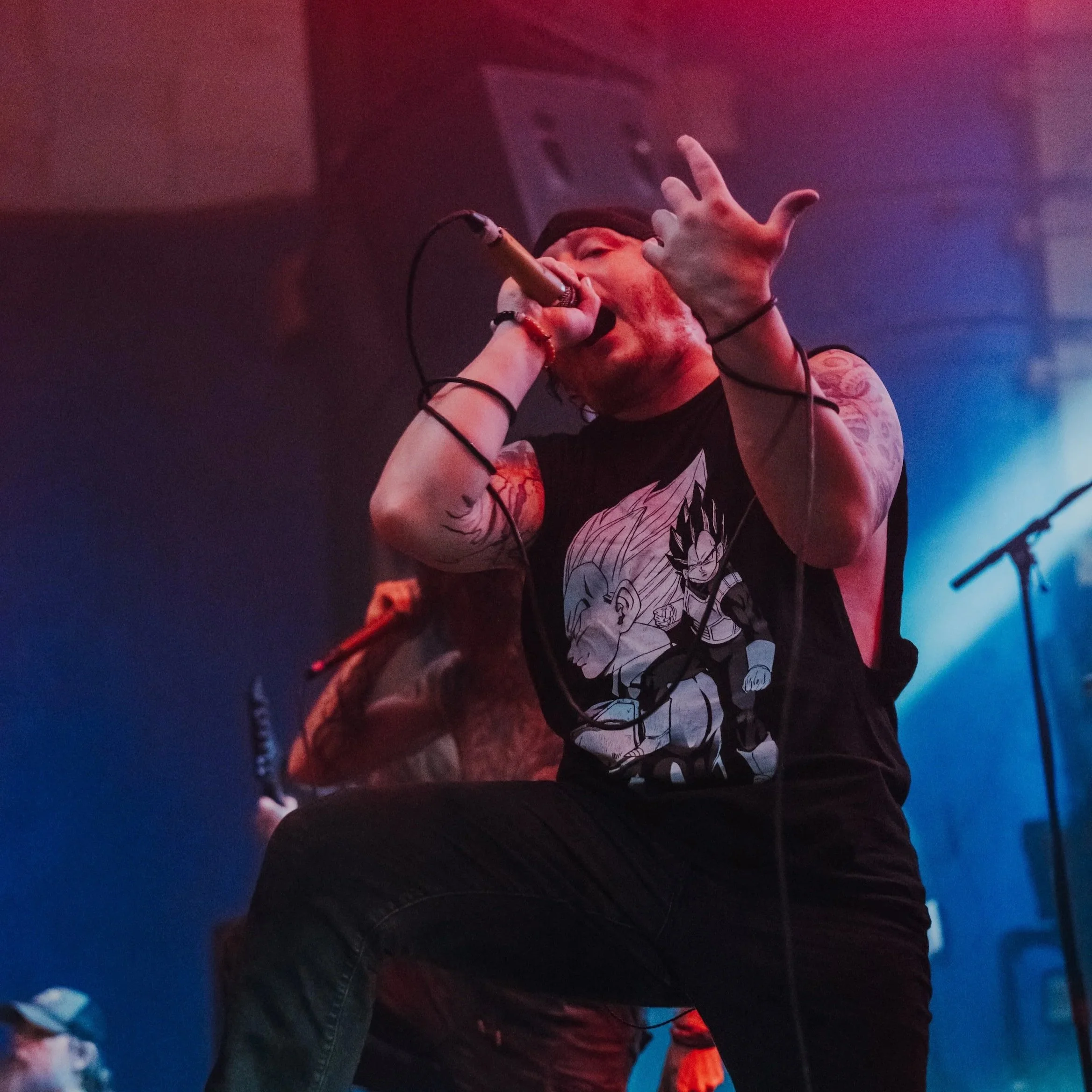 A male vocalist performs passionately with a microphone in a dimly lit concert setting, wearing a black sleeveless shirt featuring anime characters, with tattoos on his arms.