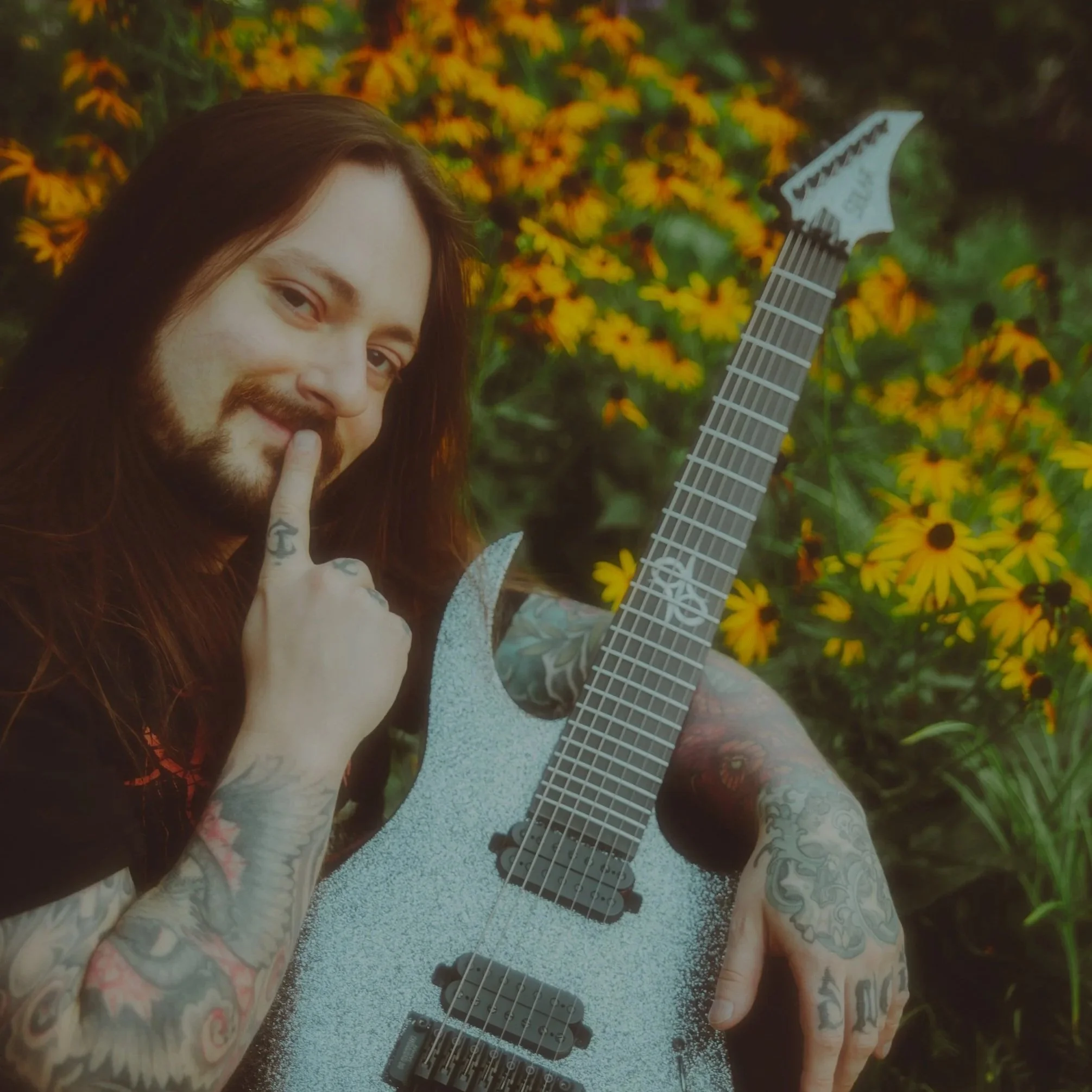 A man with long hair and tattoos holding an electric guitar with a textured finish, standing in front of yellow flowers, making a shh gesture with his finger.
