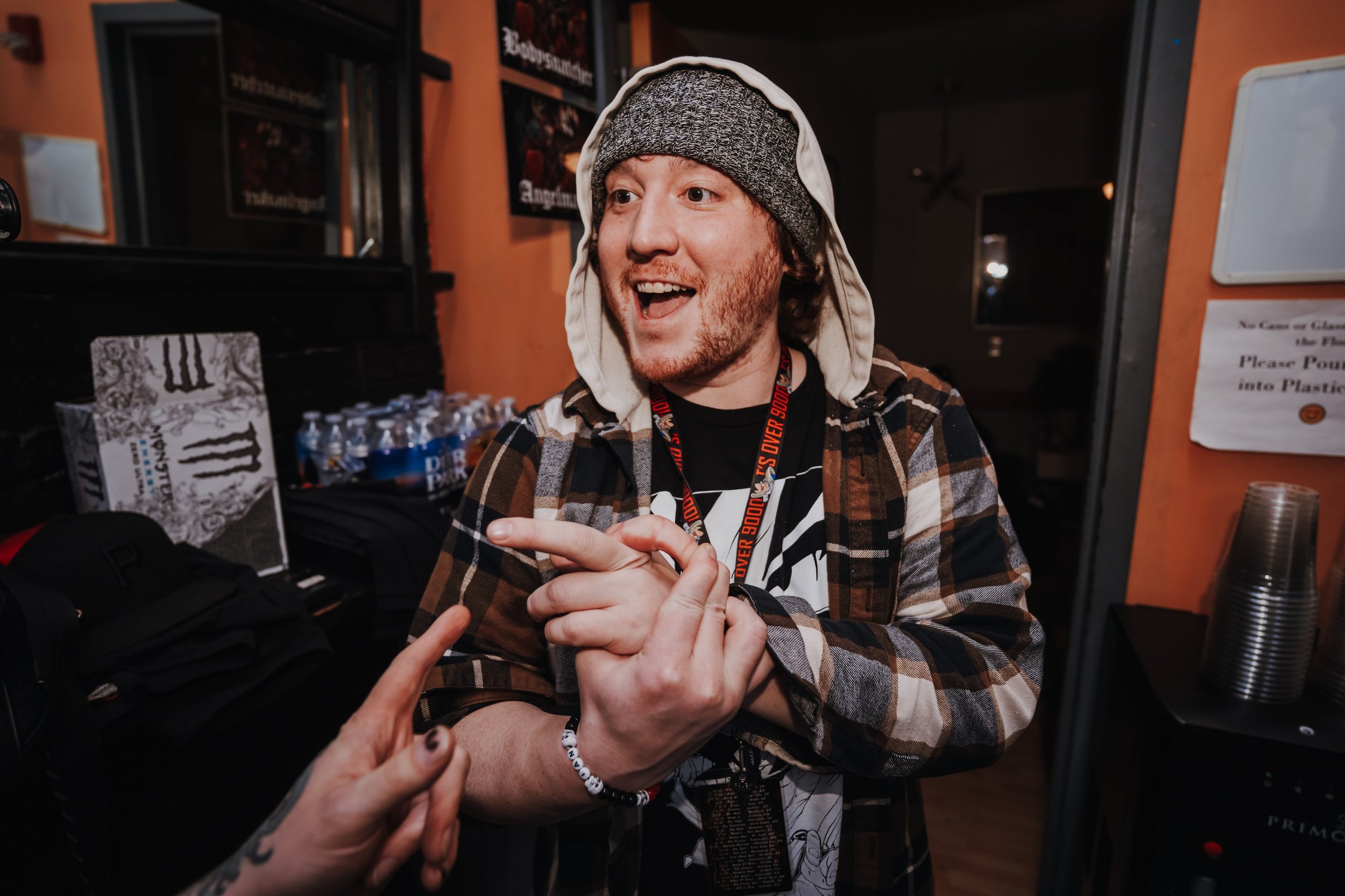 A young man with a beard, wearing a gray beanie, a hood, and a plaid jacket, smiling and gesturing with his hands while talking to someone indoors at a social event.