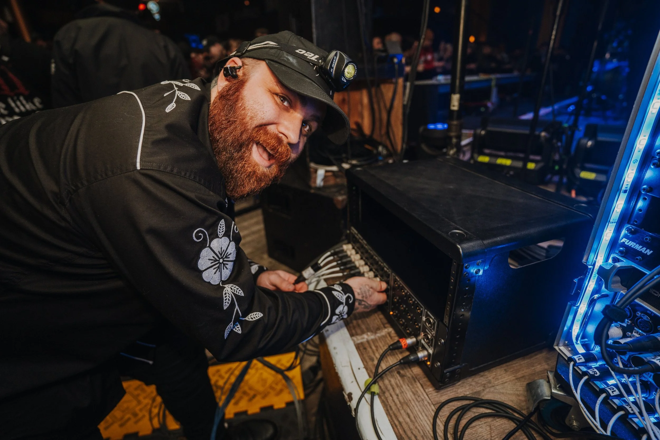A man with a beard and a black hat, smiling, working with audio equipment at a soundboard during an event.