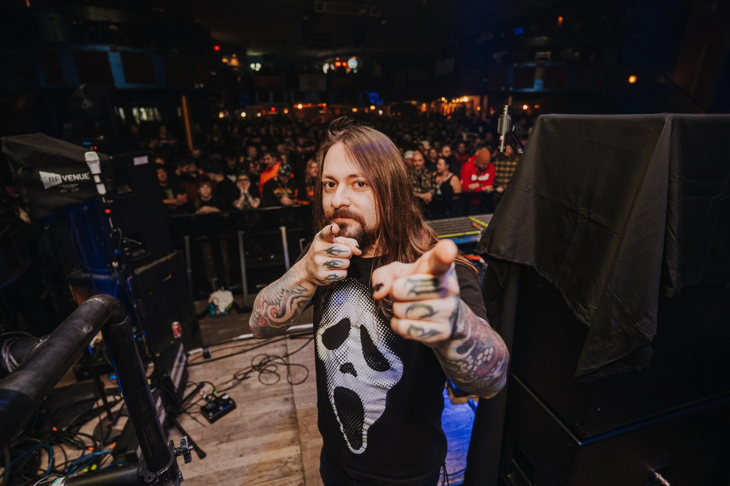 A man with long hair and tattoos is standing on a stage, pointing at the camera, with an enthusiastic crowd behind him at an indoor concert or event.