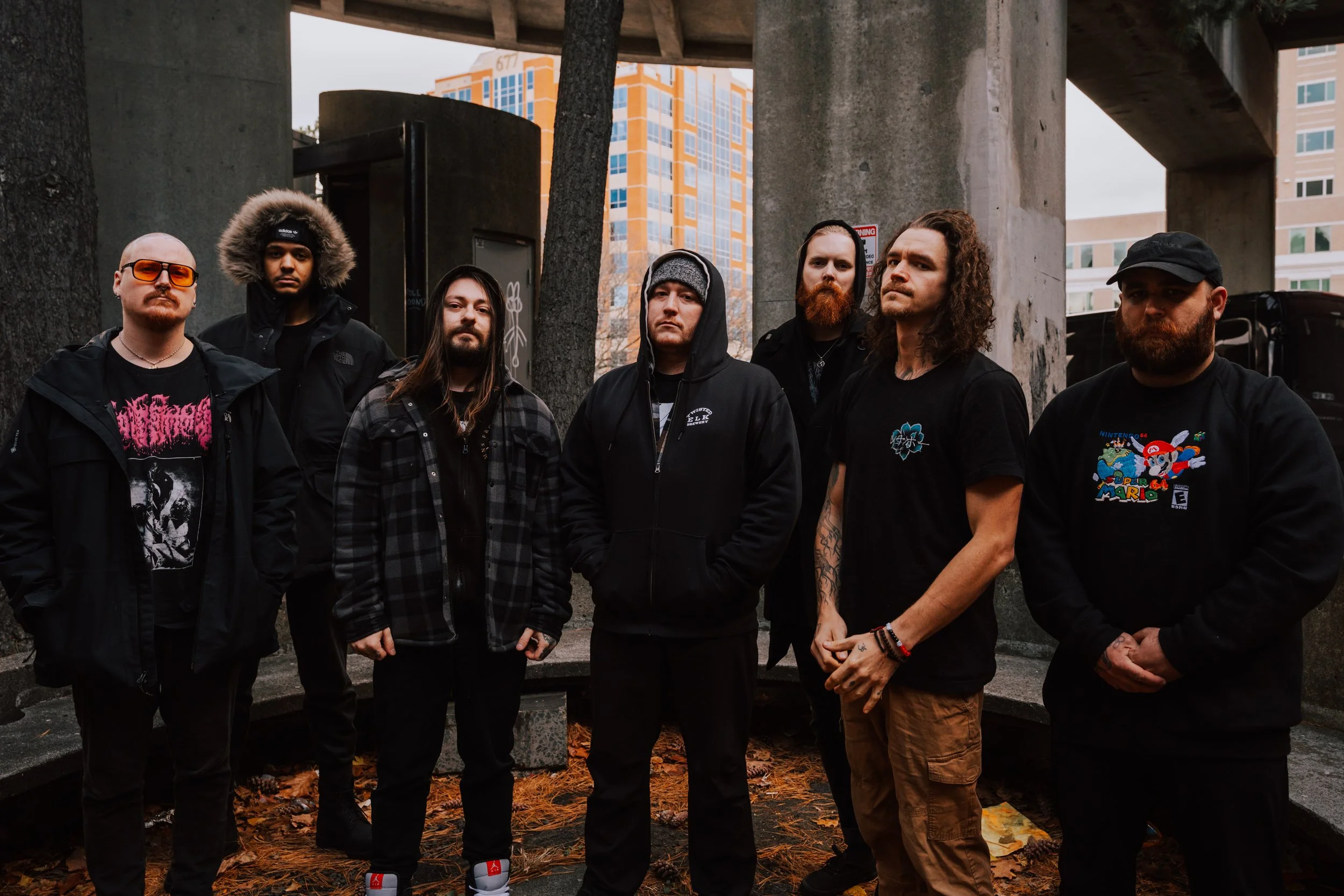 Group of seven men standing outdoors under a bridge with buildings in the background, dressed in casual and dark clothing.