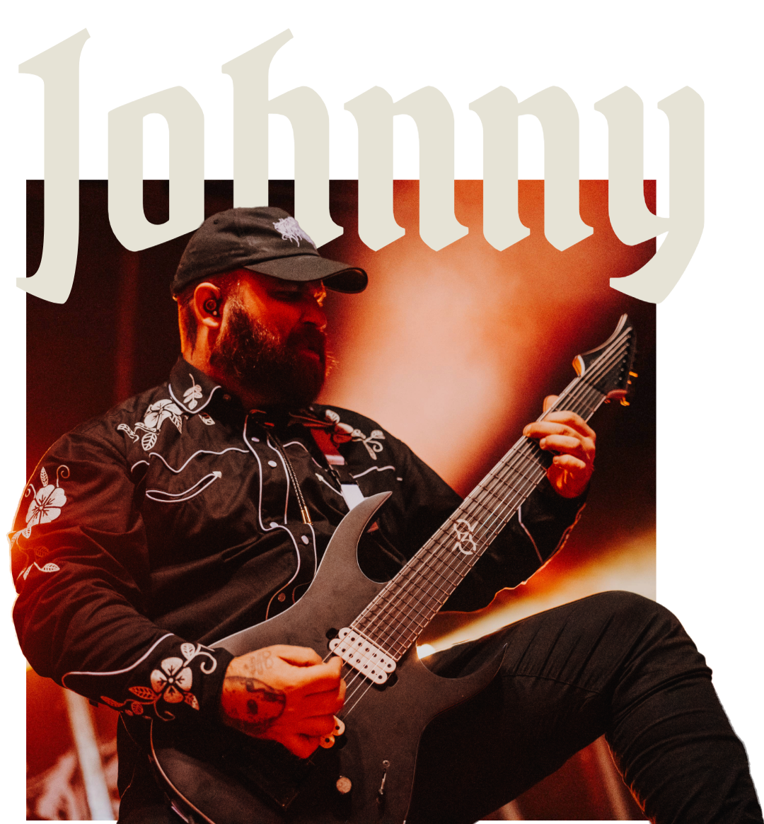 A male musician with a beard, wearing a black cap and black embroidered shirt, playing an electric guitar on stage with warm lighting. Part of the 'Jou' text is visible at the top.