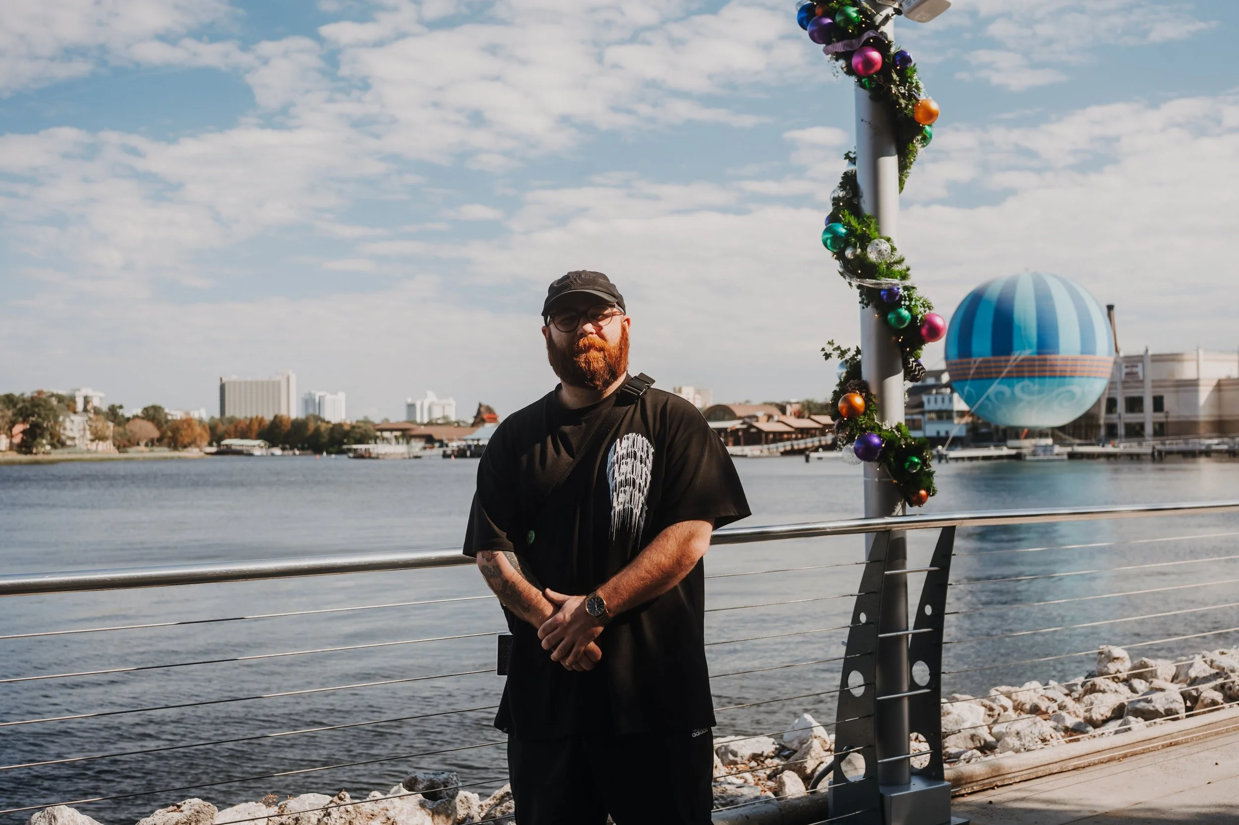 A man with a beard and glasses wearing a black t-shirt with a graphic design, standing near a body of water during daytime, with holiday decorations on a nearby pole and a hot air balloon in the background.