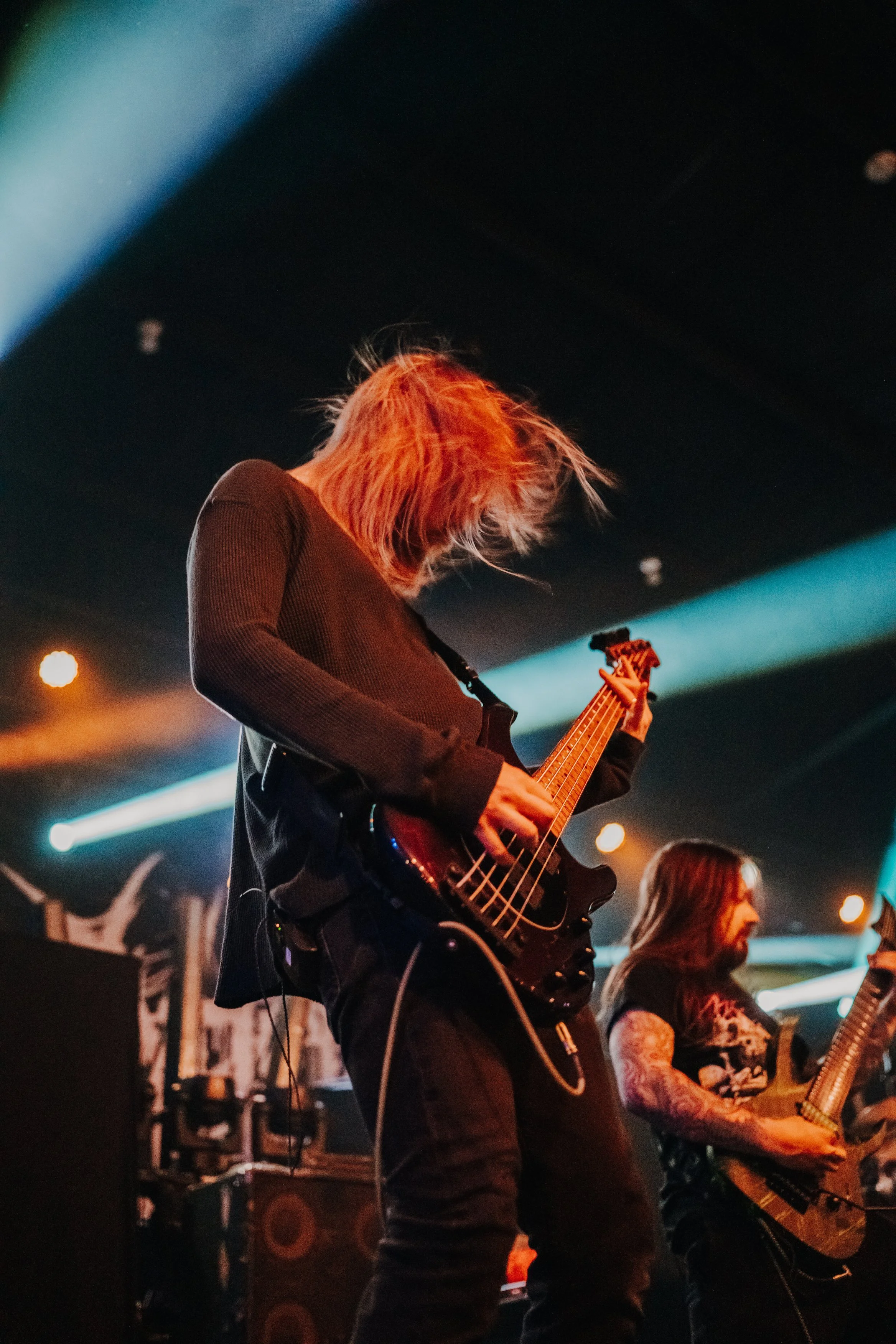 Two musicians performing on stage, one playing a bass guitar and the other playing an electric guitar, with stage lighting and equipment in the background.