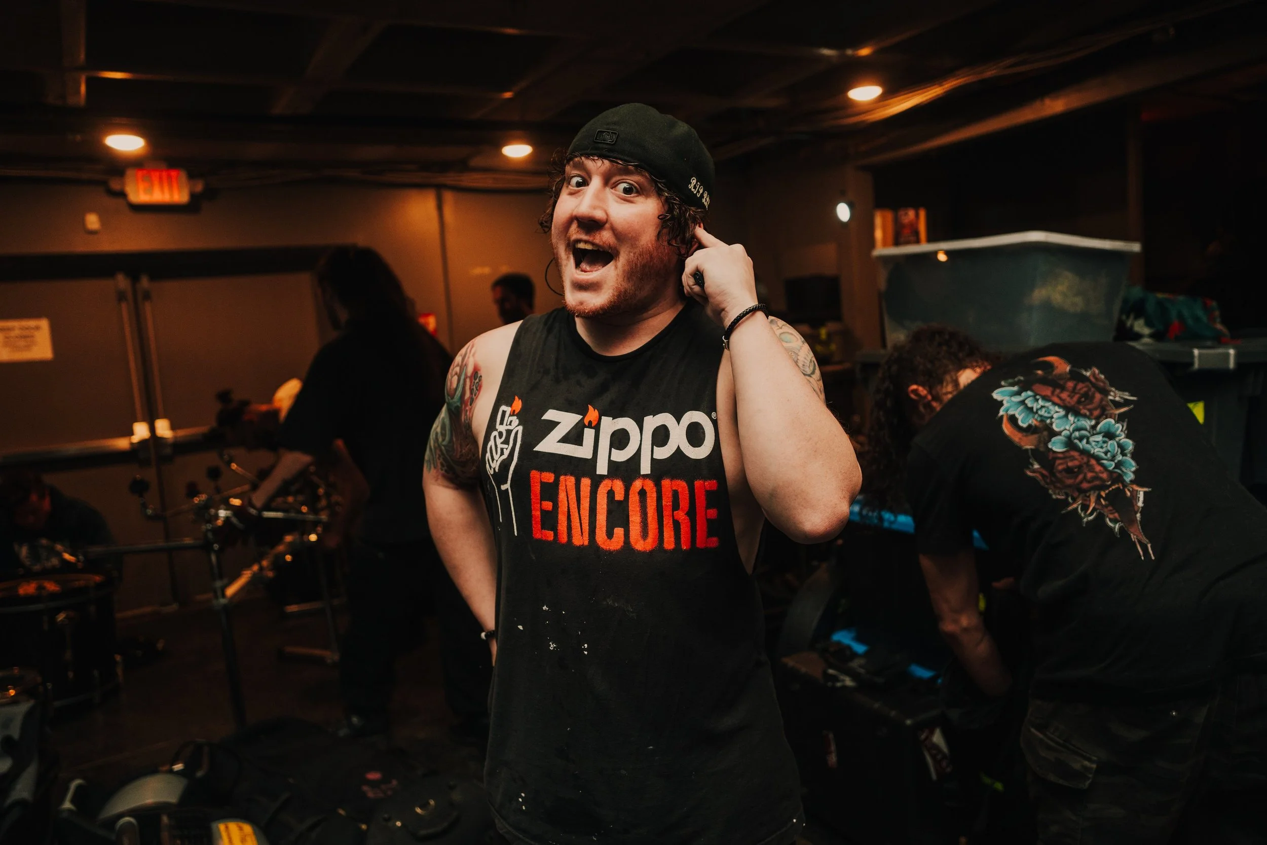 Smiling man with tattoos on arms wearing a black ZippO ENCORE sleeveless shirt and a backwards black cap, holding an earpiece, in a dimly lit room with other people around, some working on equipment.