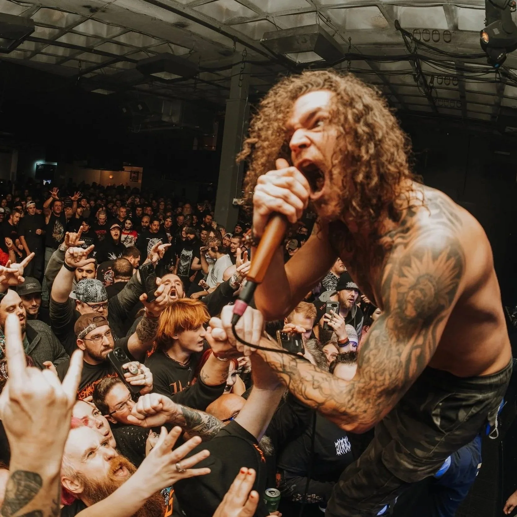 A shirtless male lead singer with curly hair and tattoos passionately singing into a microphone while crowd surfing at a concert.