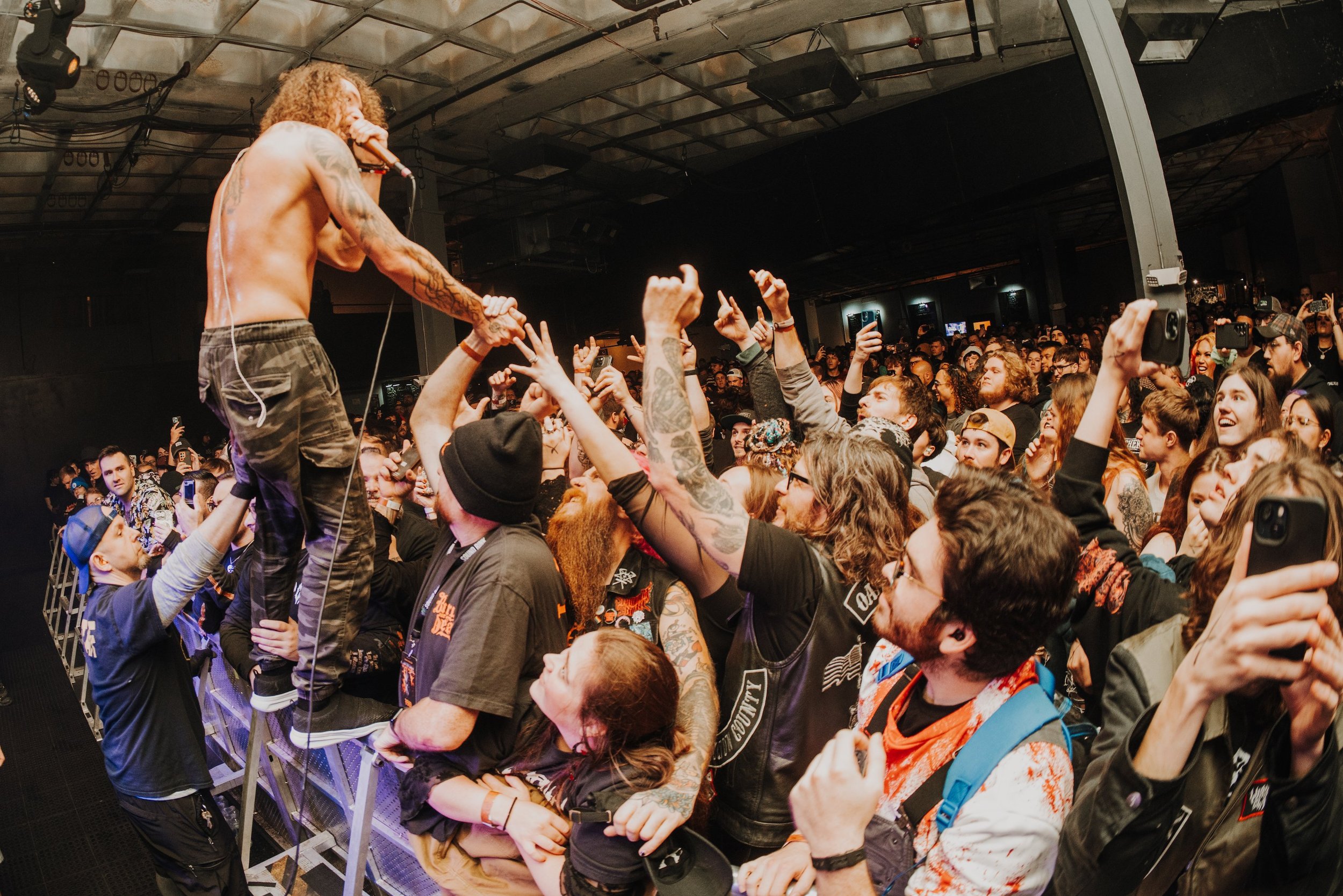 A shirtless male performer singing into a microphone while standing on the barrier surrounded by excited concertgoers reaching out to him. The crowd is diverse, with some taking pictures, and the venue has a dark industrial ceiling.